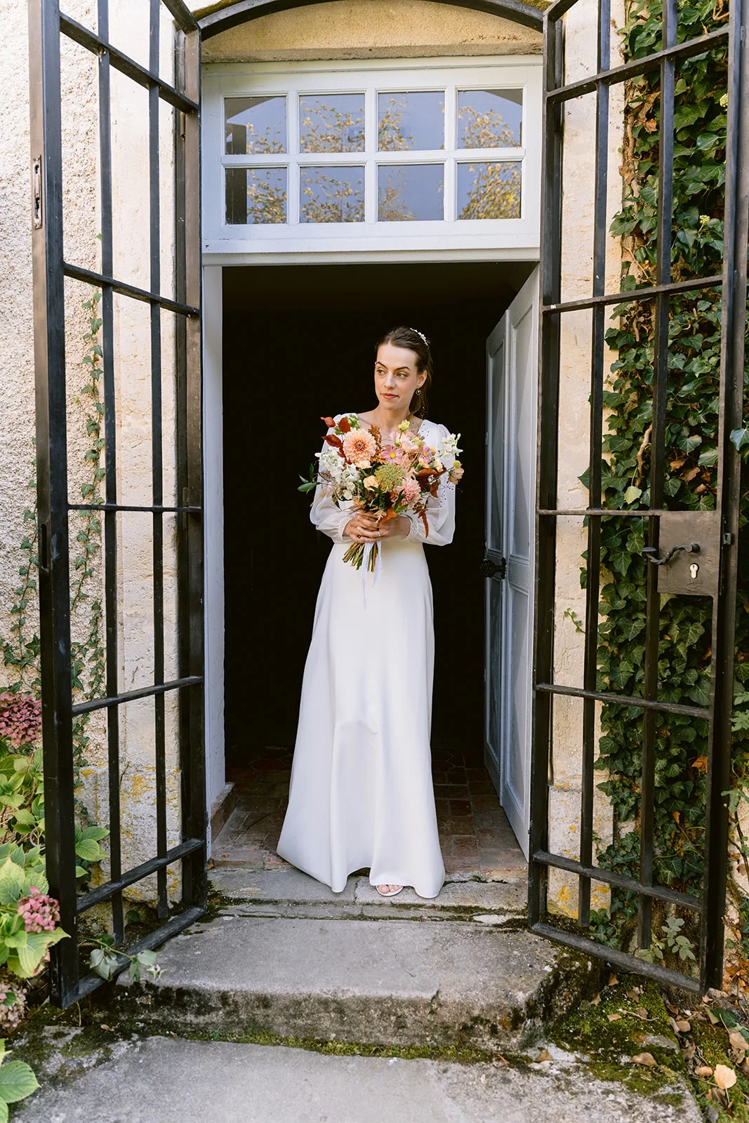 La mariée pose avec son bouquet sur le seuil d’une porte, entre deux grilles ouvertes.