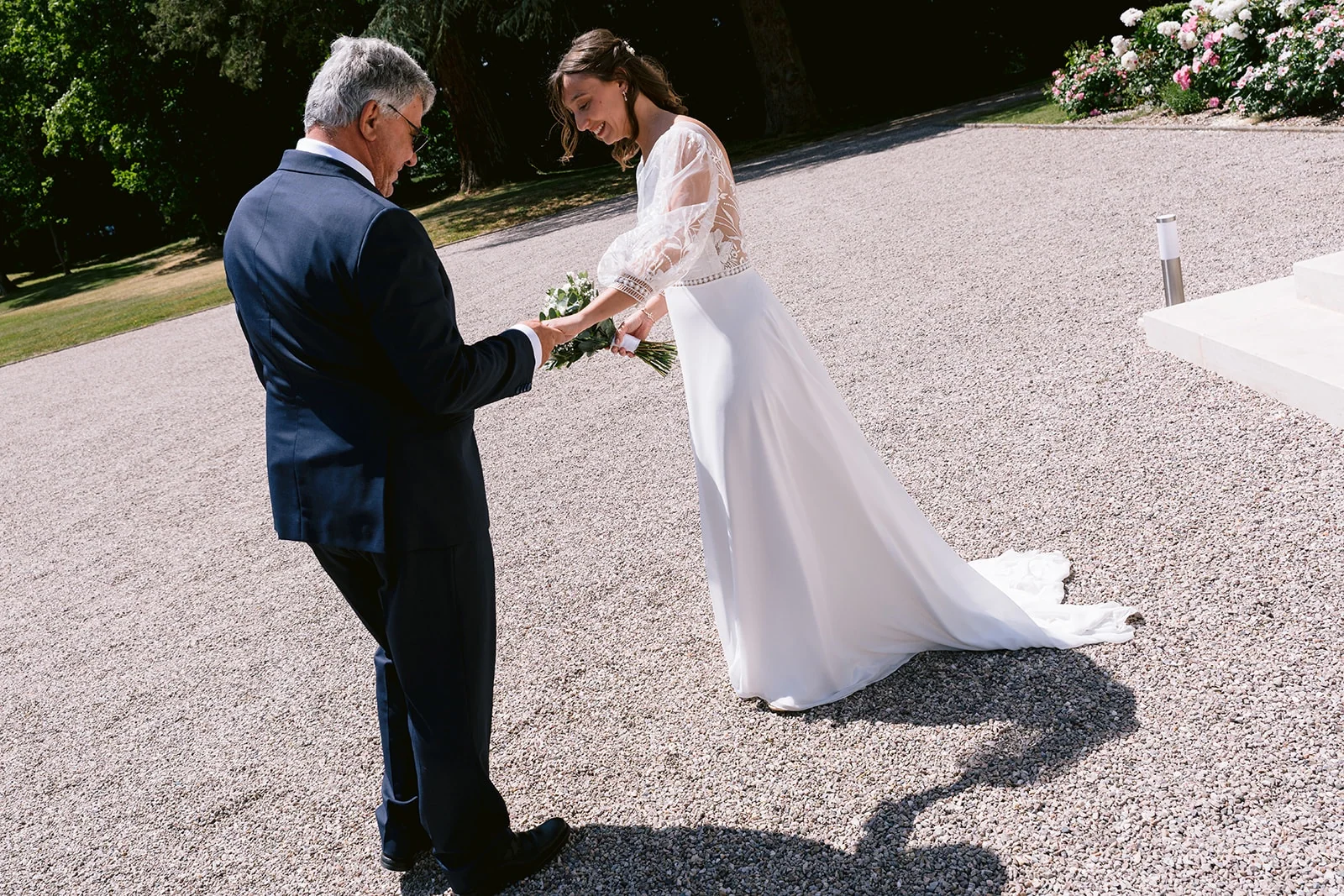 La mariée tient la main d’un homme en costume bleu dans une cour ensoleillée, son bouquet dans l’autre main.