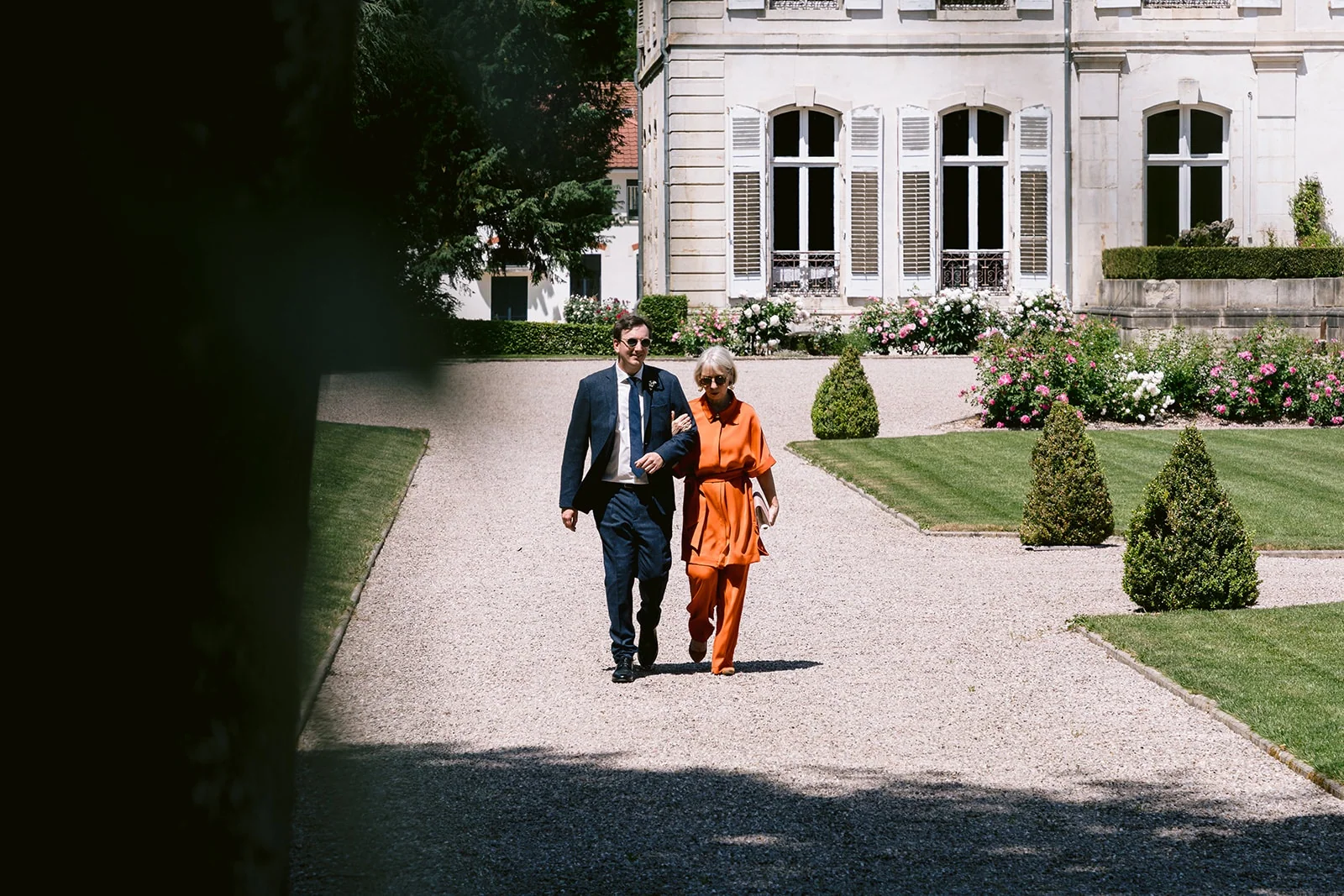 Le marié avance au bras d’une femme en tenue orange sur l’allée qui relie la demeure au jardin.