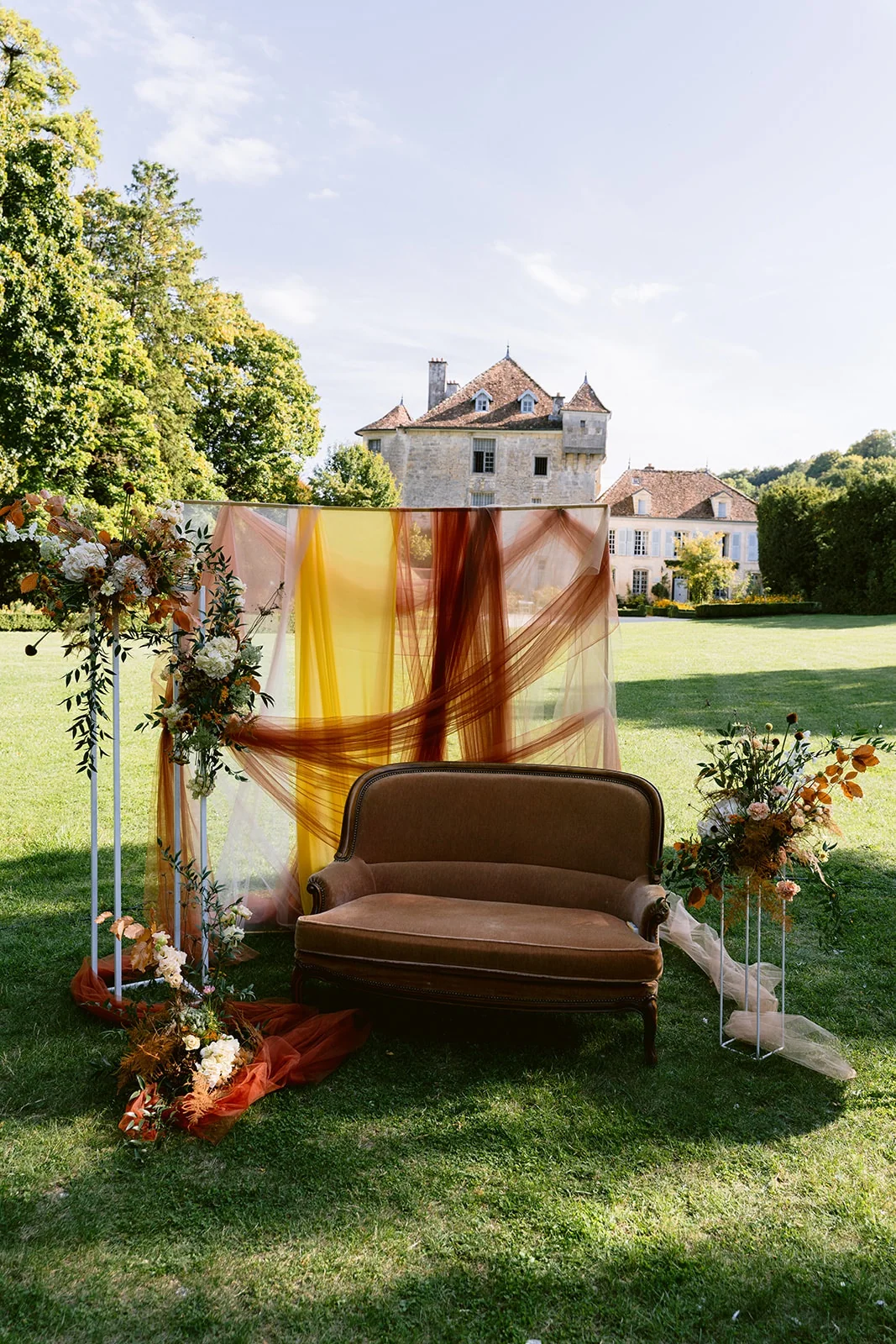 Une banquette et des fleurs composent un décor de cérémonie aux drapés orangés, face à une demeure en pierre.