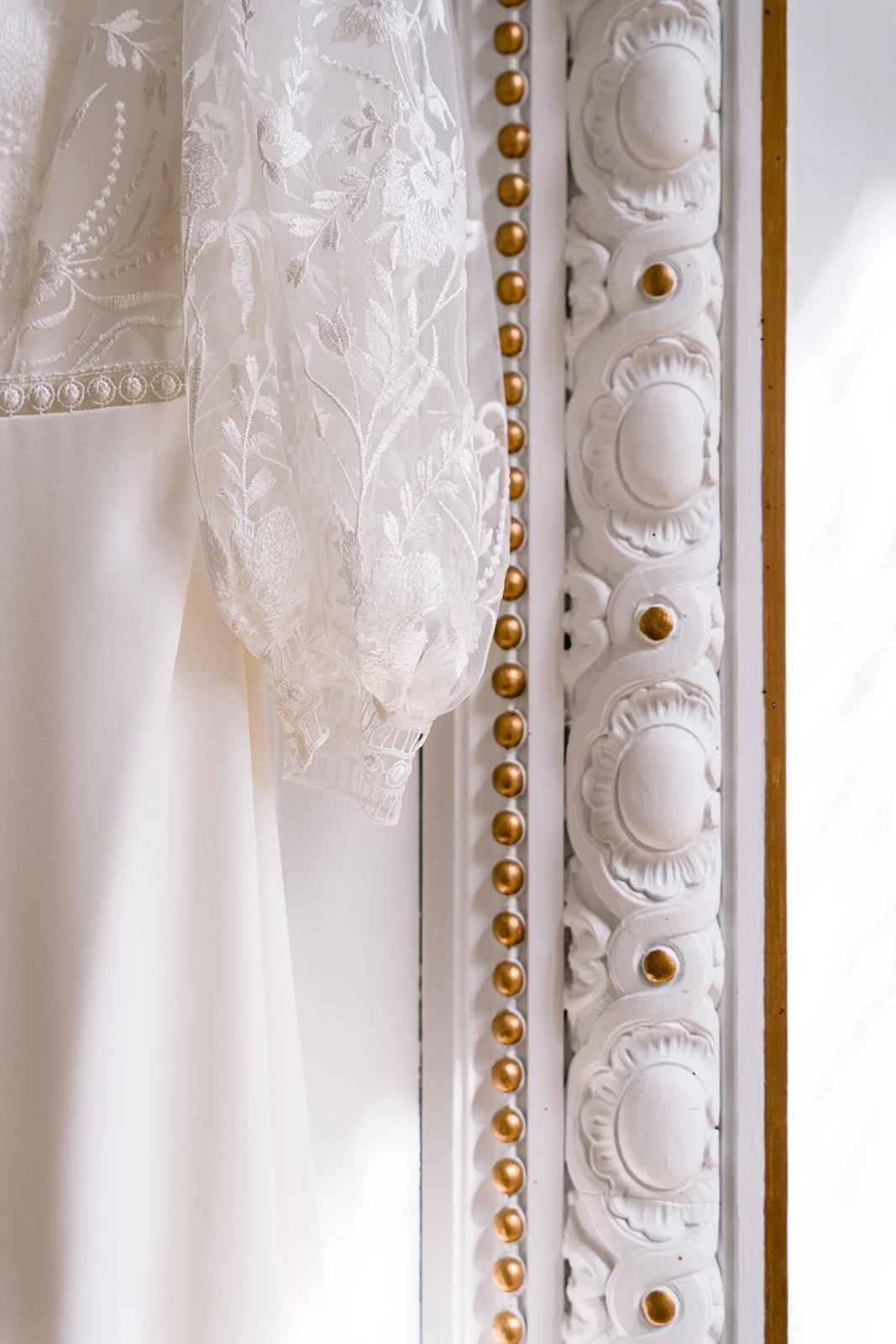 Gros plan sur la manche en dentelle florale d’une robe de mariée, près d’un encadrement blanc aux détails dorés.