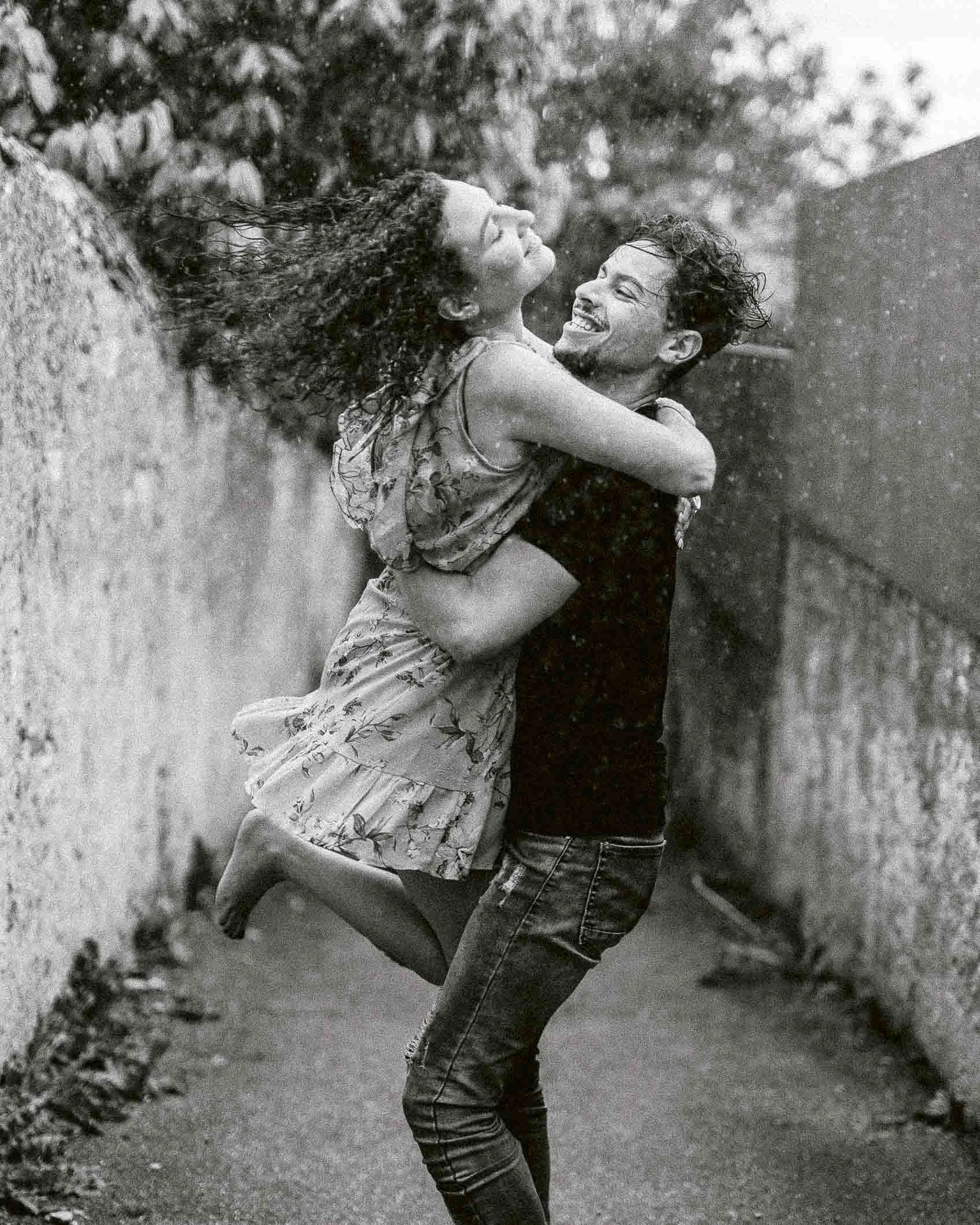 En noir et blanc, un homme soulève sa compagne qui rit, dans une allée bordée d’un mur.