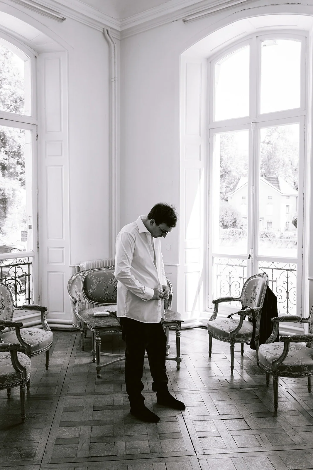 En noir et blanc, le marié ajuste sa tenue devant les hautes fenêtres d’un salon.