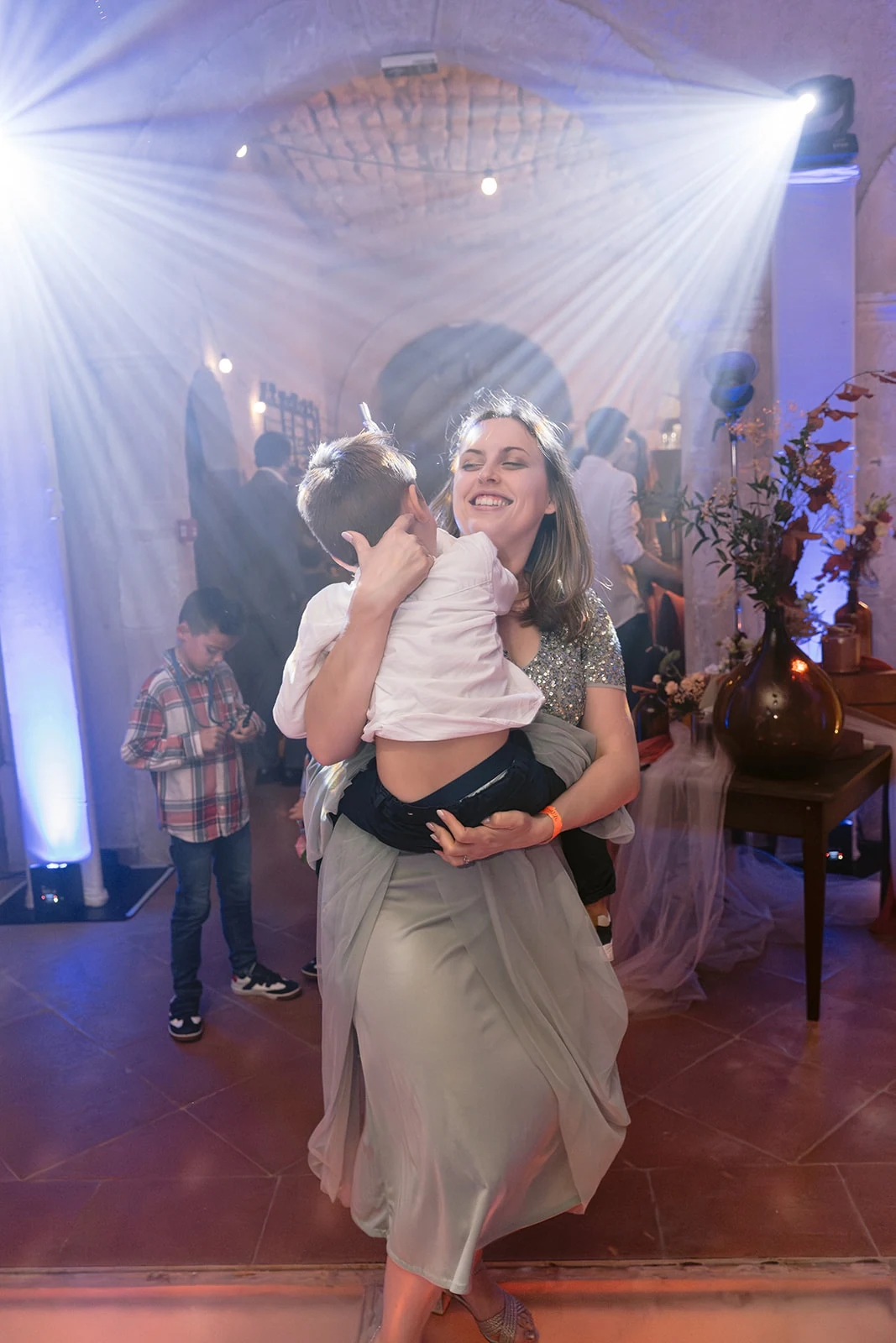Une femme souriante tient un enfant dans ses bras sous les faisceaux lumineux de la piste de danse.