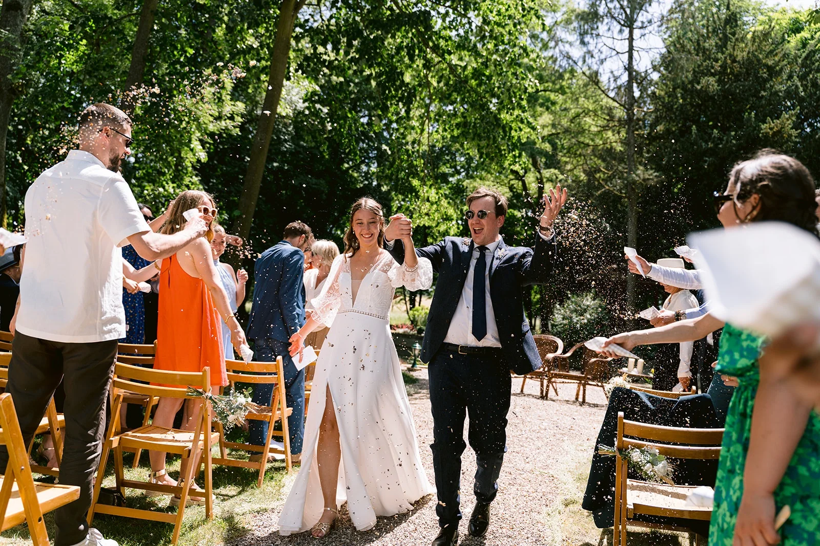 Les mariés traversent l’allée de la cérémonie parmi les invités qui les applaudissent, le marié levant un bras.