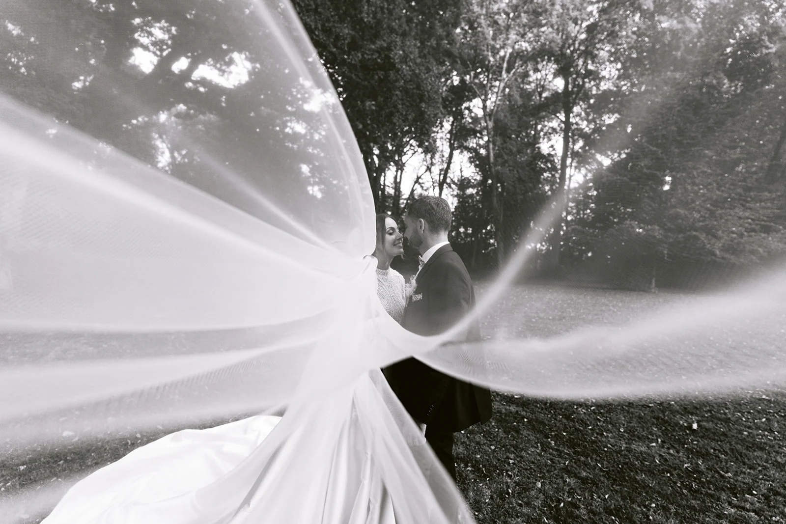 En noir et blanc, le voile de la mariée se déploie autour du couple dans le parc.