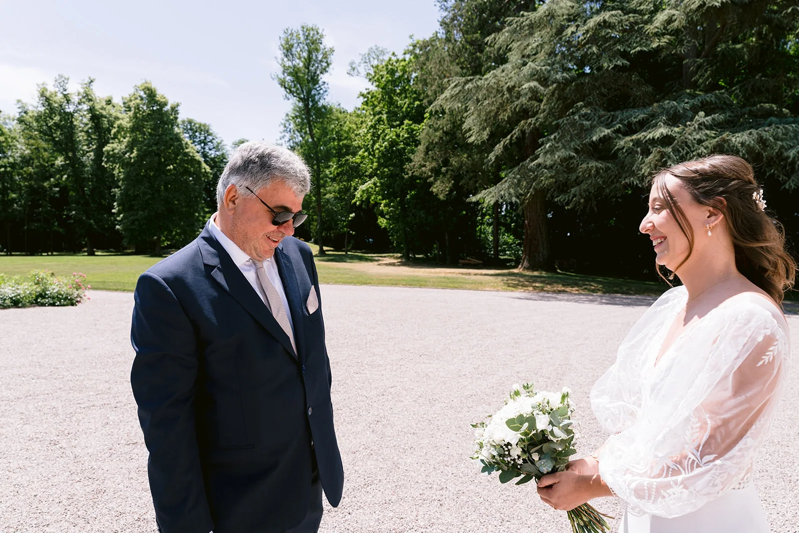 La mariée sourit à un homme aux cheveux gris en costume bleu, dans une cour ensoleillée bordée d’arbres.
