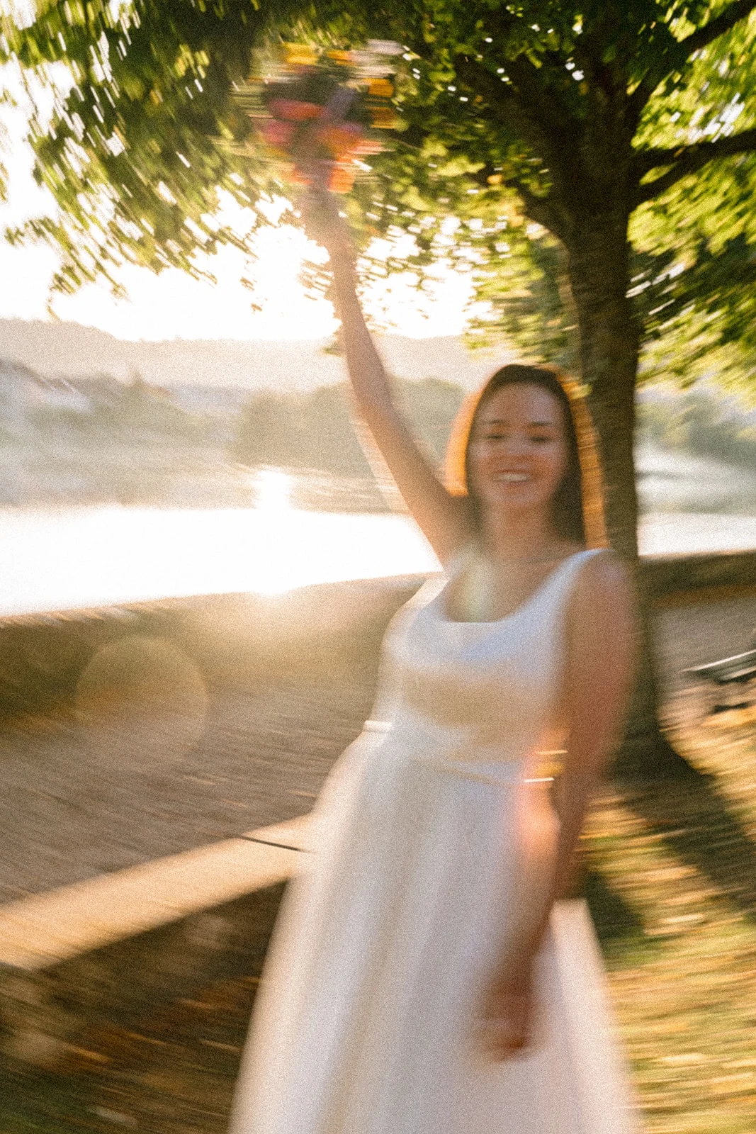 Une mariée lève le bras au bord de l’eau, dans un flou de mouvement au soleil couchant.