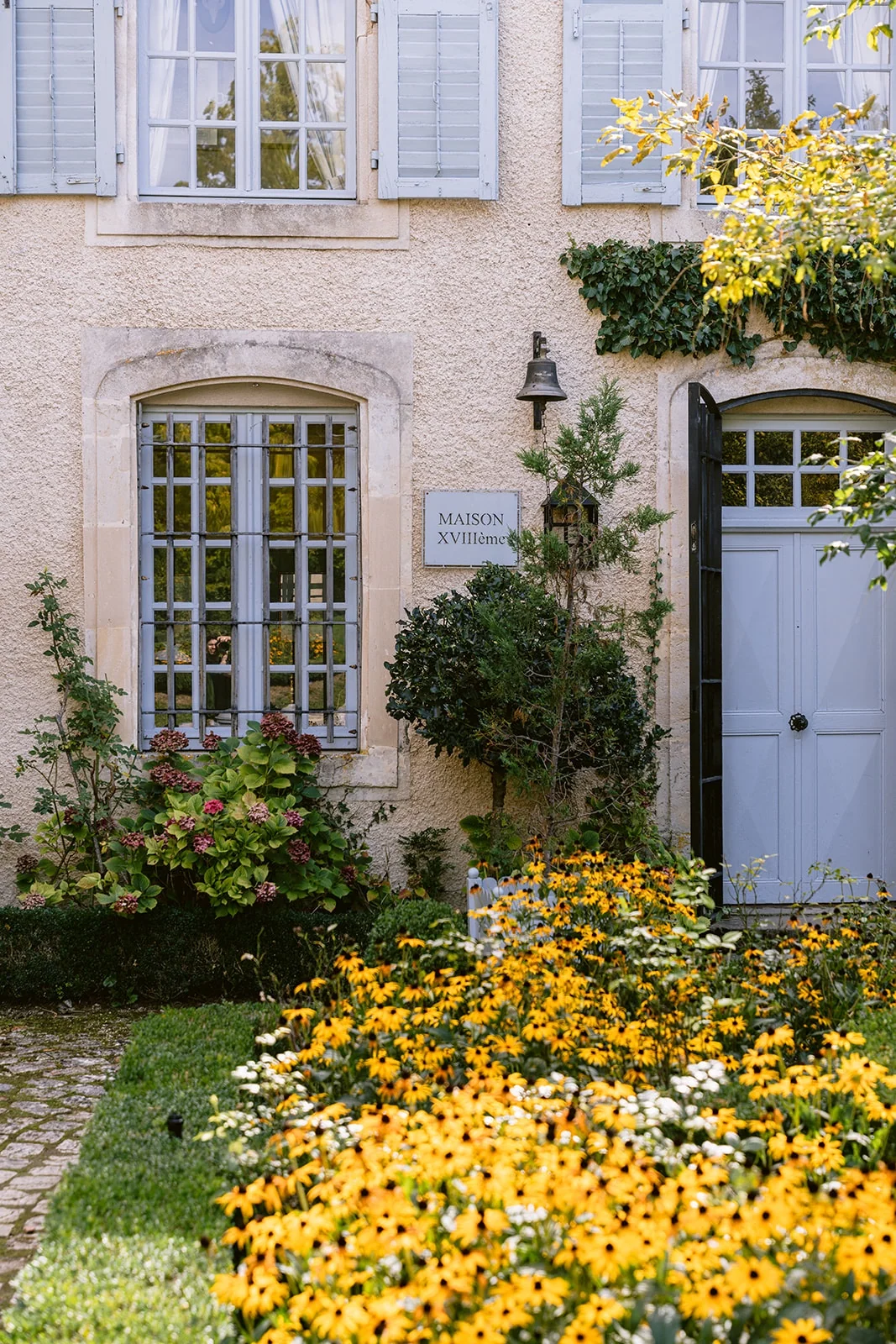 Des fleurs jaunes bordent la façade claire d’une maison aux volets et à la porte bleu pâle.