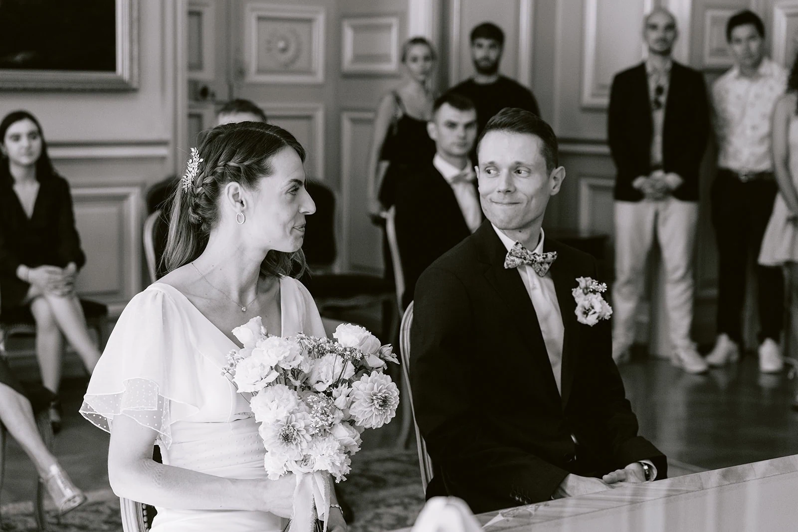 En noir et blanc, les mariés échangent un regard pendant la cérémonie, devant leurs invités.