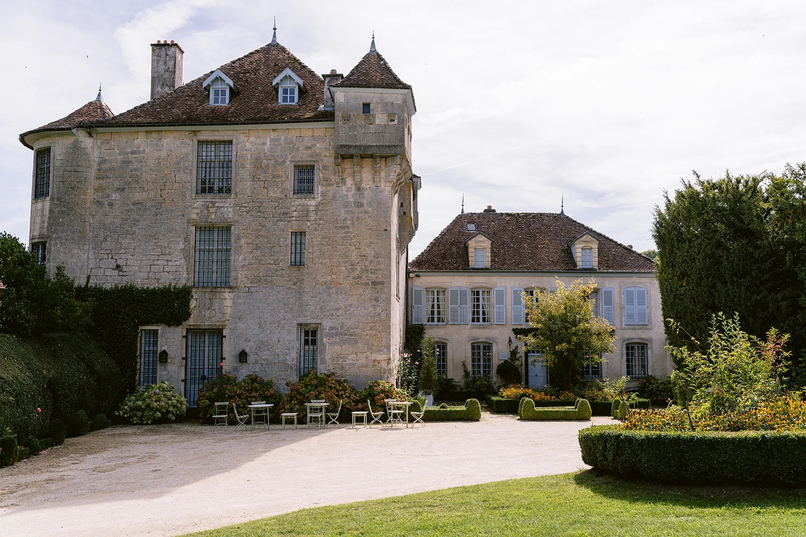 La façade d’un château de pierre avec une tour carrée et des volets clairs, devant une cour et un jardin.