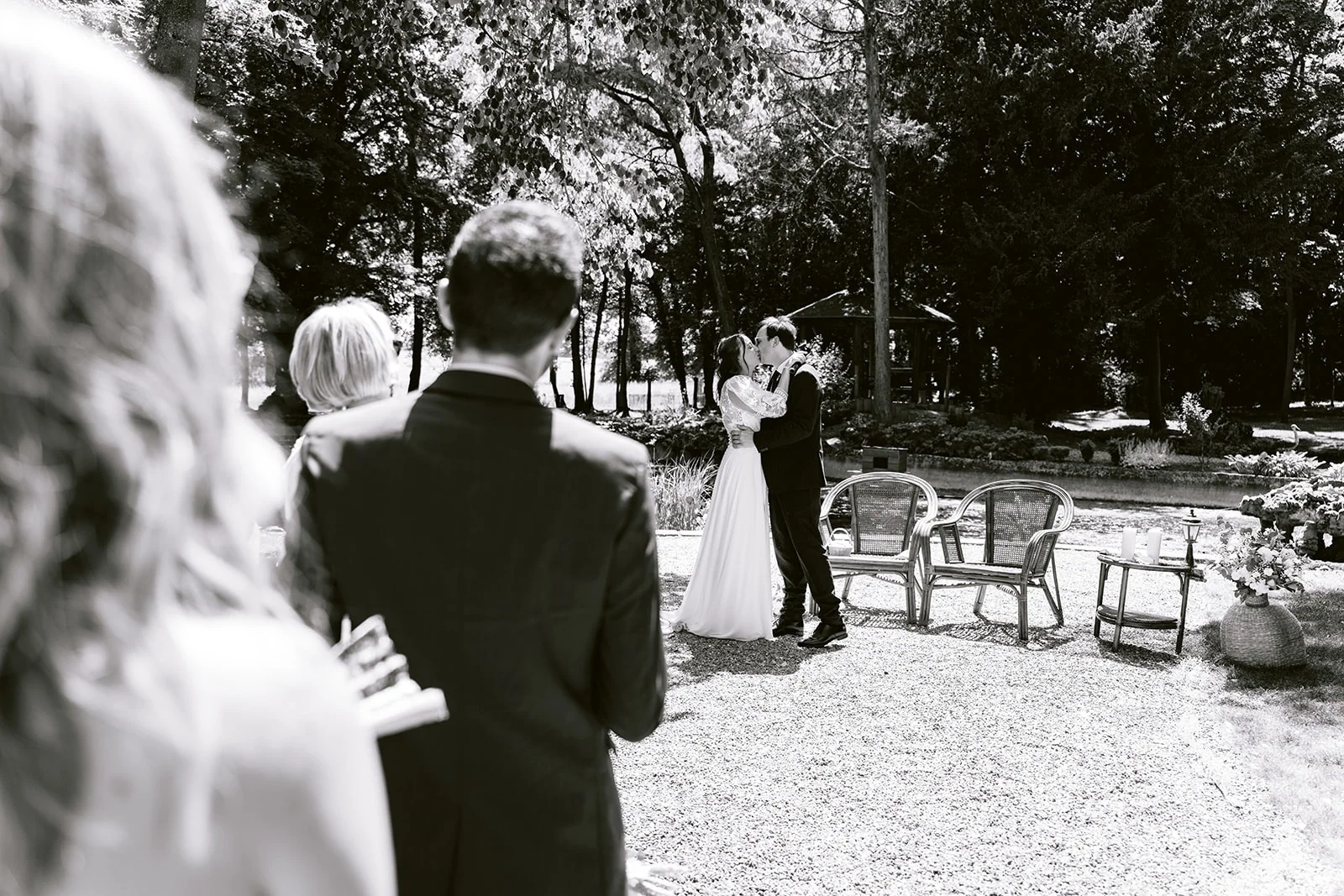 En noir et blanc, les mariés s’embrassent sous les arbres devant les invités, près de deux fauteuils en rotin.