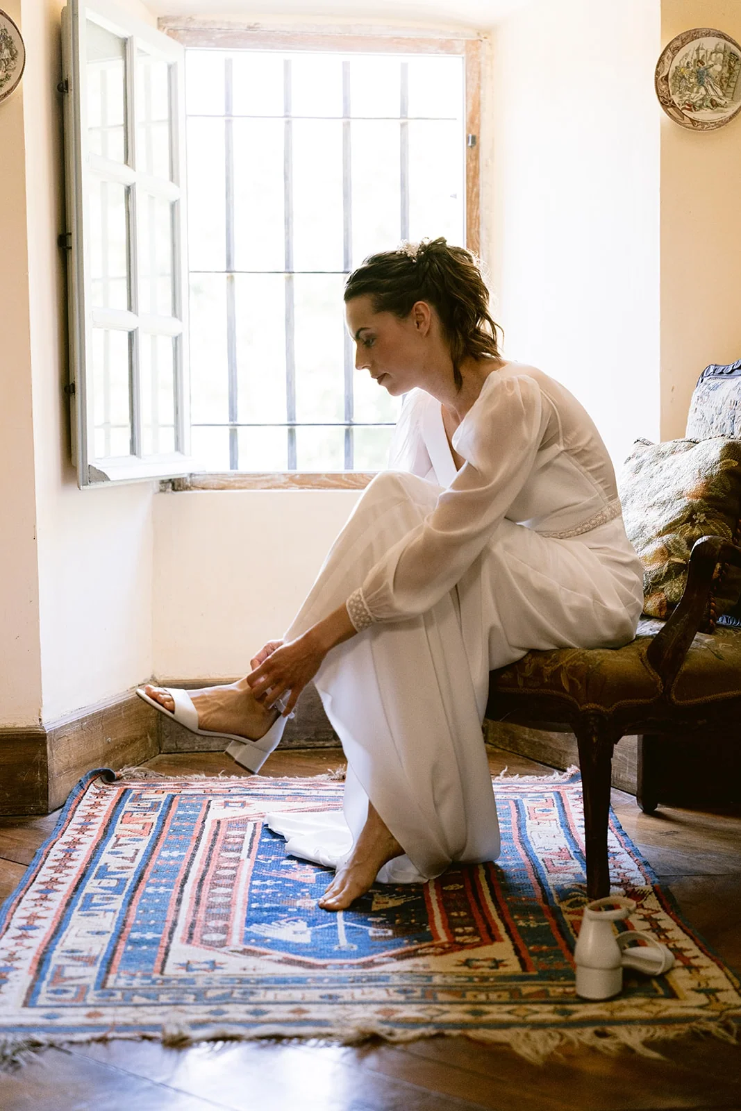 La mariée ajuste la bride de sa sandale blanche, assise au bord d’un fauteuil près d’une fenêtre.