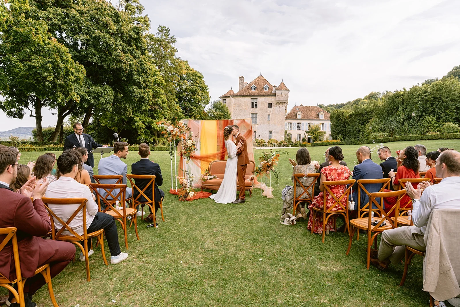 Les mariés s’embrassent devant le décor de cérémonie, sous les applaudissements des invités assis dans le jardin.