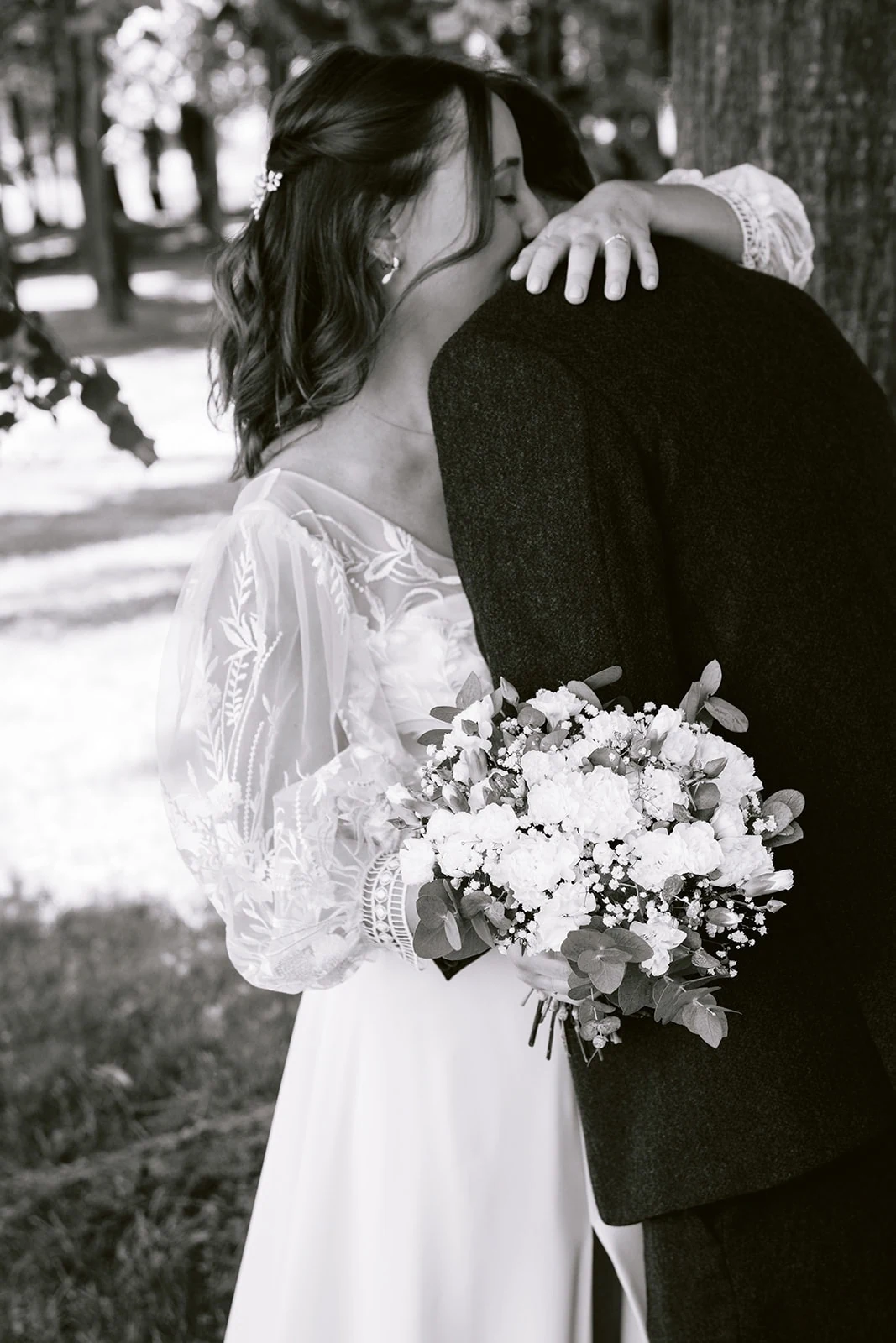 En noir et blanc, la mariée enlace le marié, une main sur son épaule et son bouquet de fleurs blanches dans le dos.