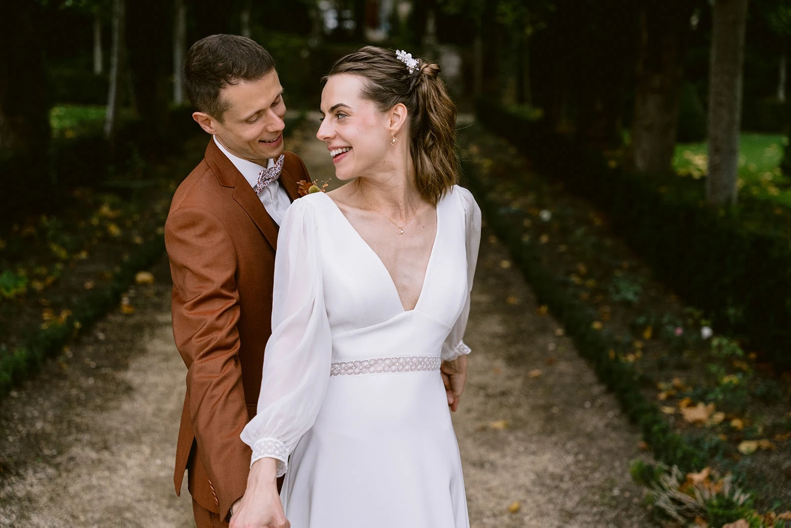 Le marié enlace la mariée par-derrière tandis qu’ils se regardent en souriant, dans une allée bordée d’arbres.