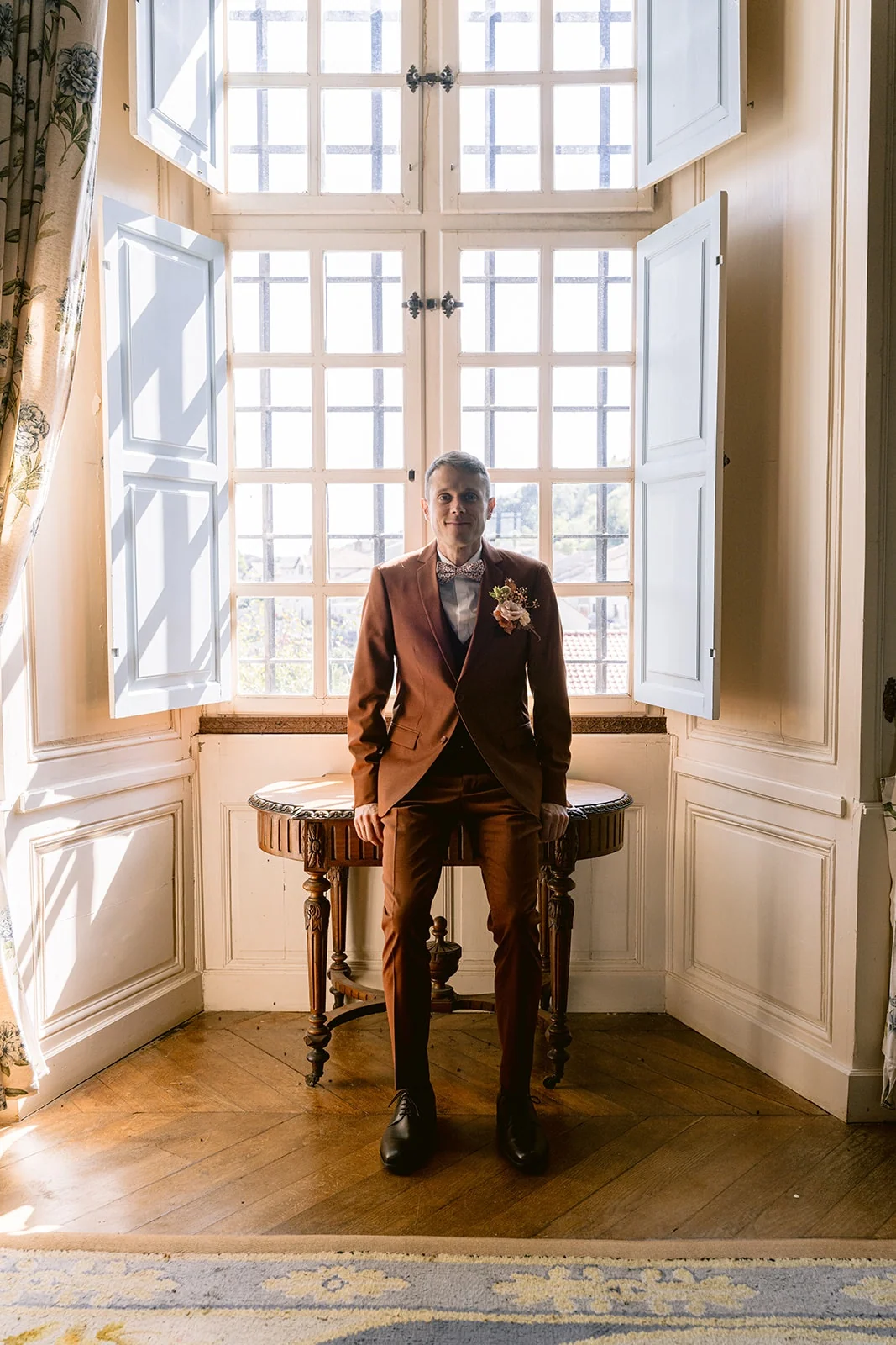 Le marié en costume brun pose devant une haute fenêtre ouverte, les mains posées sur une petite table.