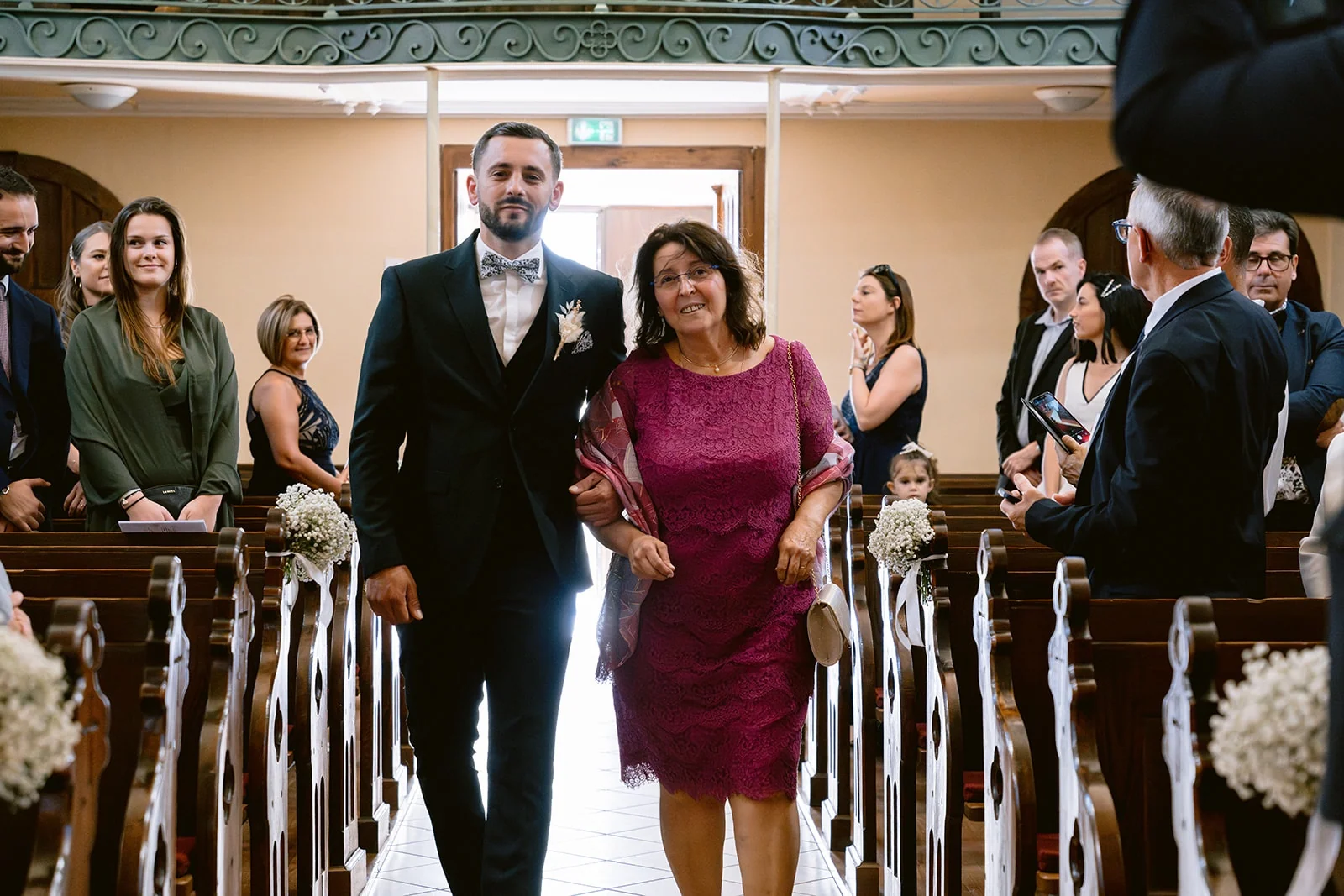 Le marié avance dans l’allée de l’église au bras d’une proche, parmi les invités.