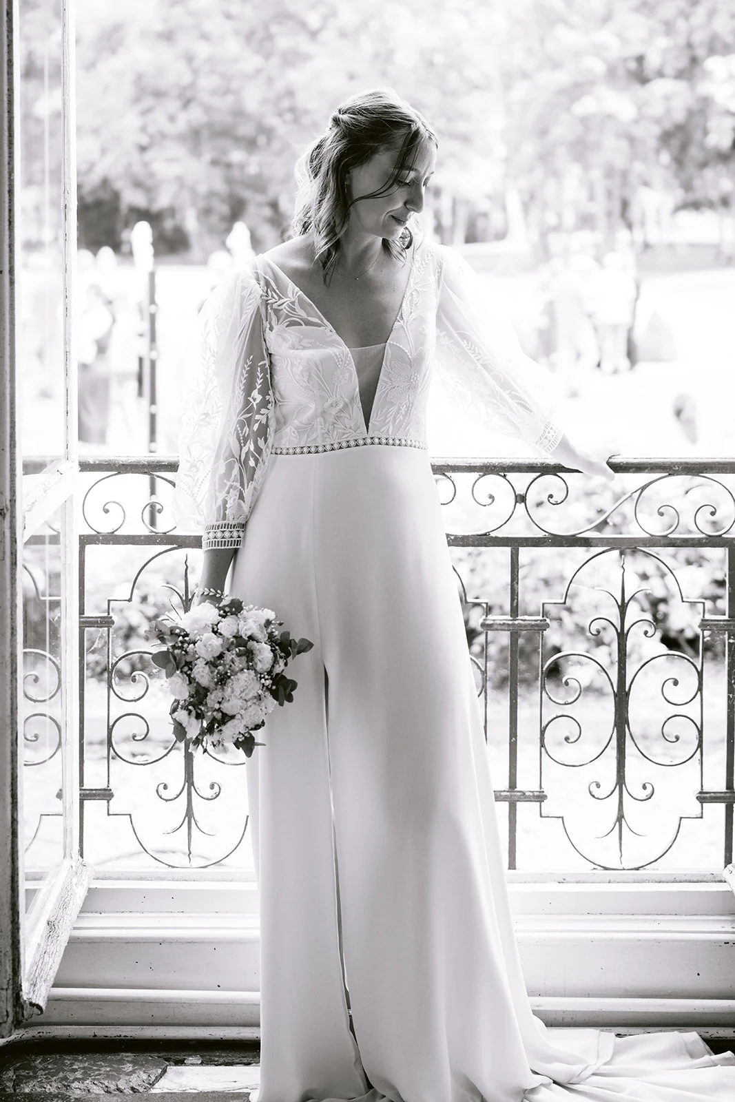 En noir et blanc, la mariée pose près d’une fenêtre ouverte sur un balcon, bouquet à la main et regard tourné au loin.
