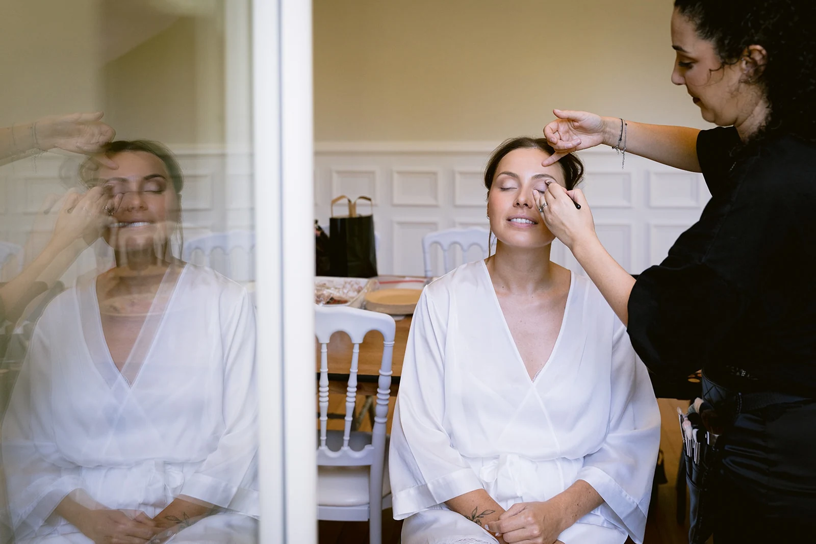 La mariée ferme les yeux pendant son maquillage, son reflet visible dans la vitre.