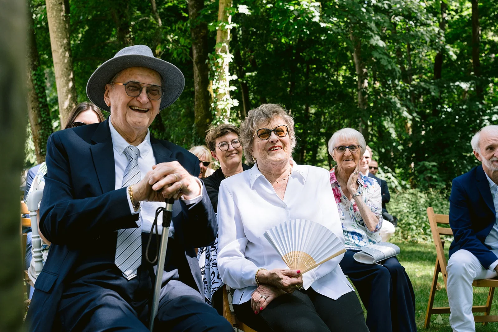 Des invités assis sous les arbres sourient, dont un homme coiffé d’un chapeau et une femme tenant un éventail blanc.
