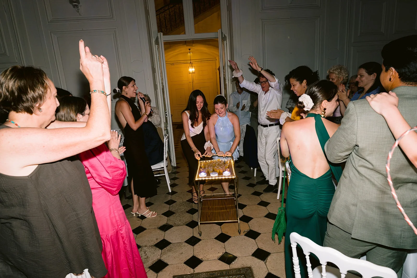 Deux femmes présentent un gâteau garni de bougies, entourées d’invités qui applaudissent et lèvent les bras.