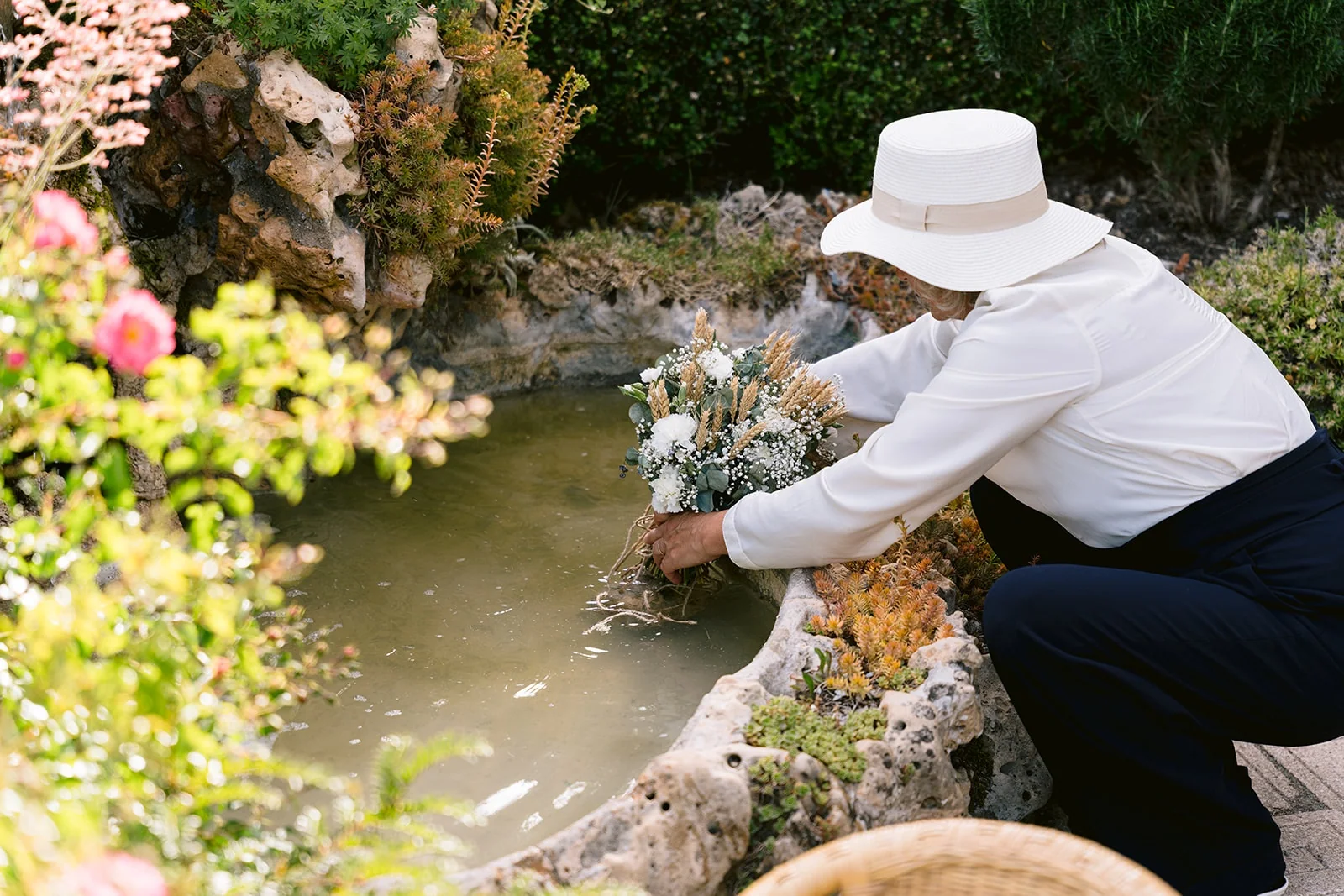 Une personne en chapeau blanc plonge les tiges d’un bouquet dans un bassin de jardin bordé de pierres.