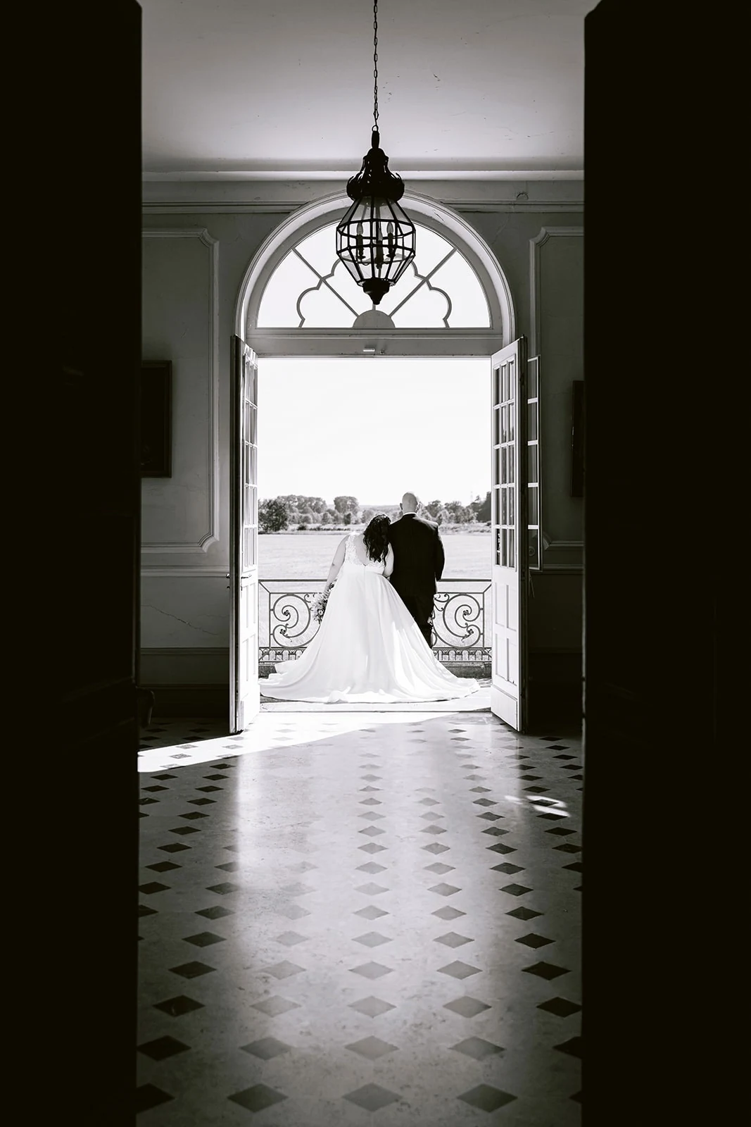 En noir et blanc, les mariés s’embrassent sur un balcon, vus depuis un hall au sol à damier.