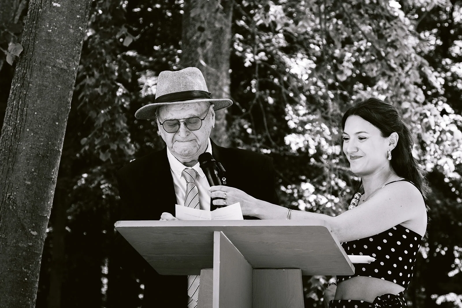 En noir et blanc, un homme coiffé d’un chapeau lit au pupitre tandis qu’une femme lui tient un microphone.