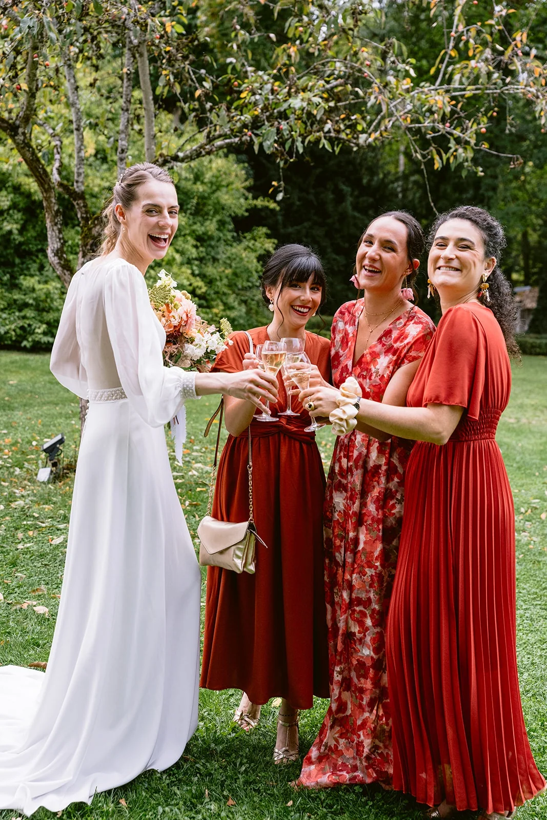 La mariée et trois femmes en robes rouges rient ensemble, leurs verres réunis dans le jardin.