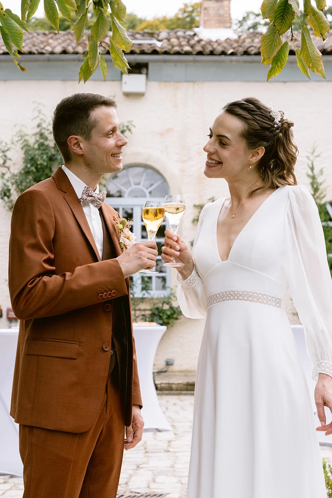 Les mariés se regardent en souriant, une flûte à la main, dans une cour pavée.