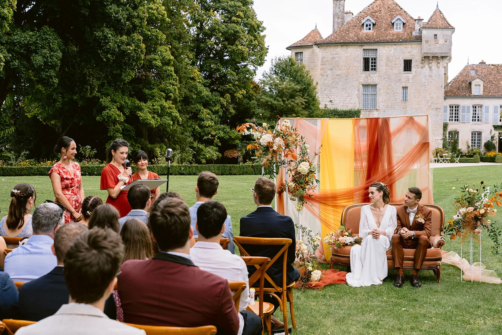 Les mariés sont assis sur une banquette devant les invités, dans un décor de fleurs et de drapés orangés.
