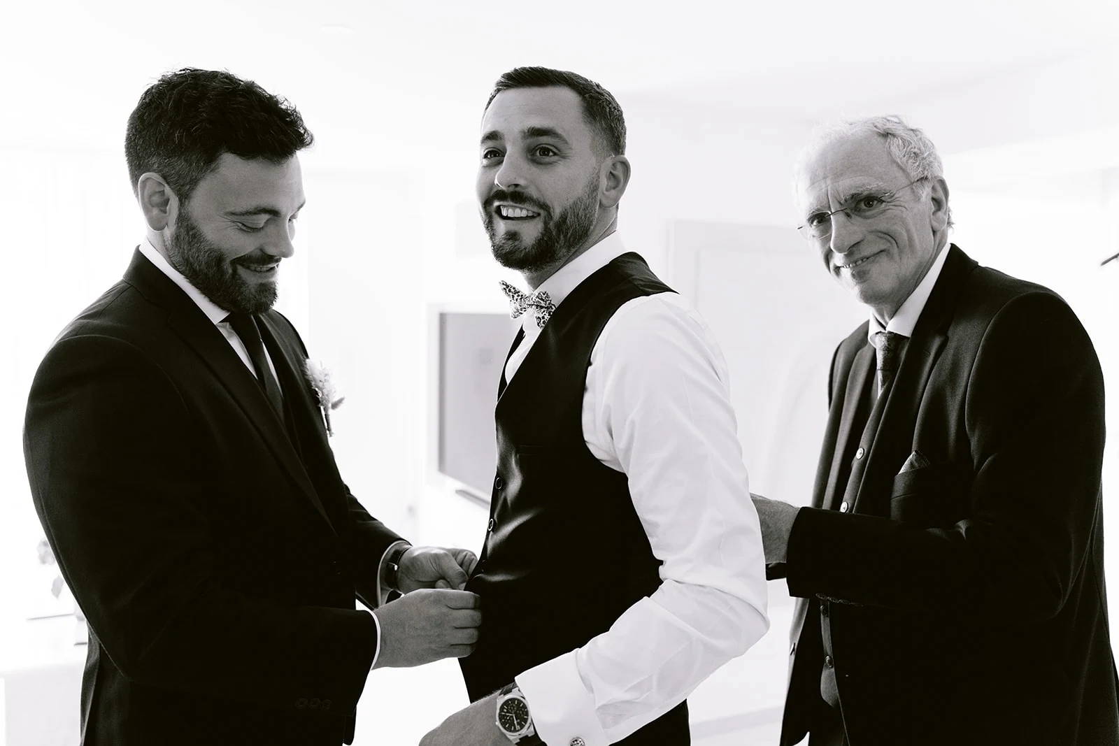 En noir et blanc, le marié en gilet rit avec deux proches pendant l’habillage.