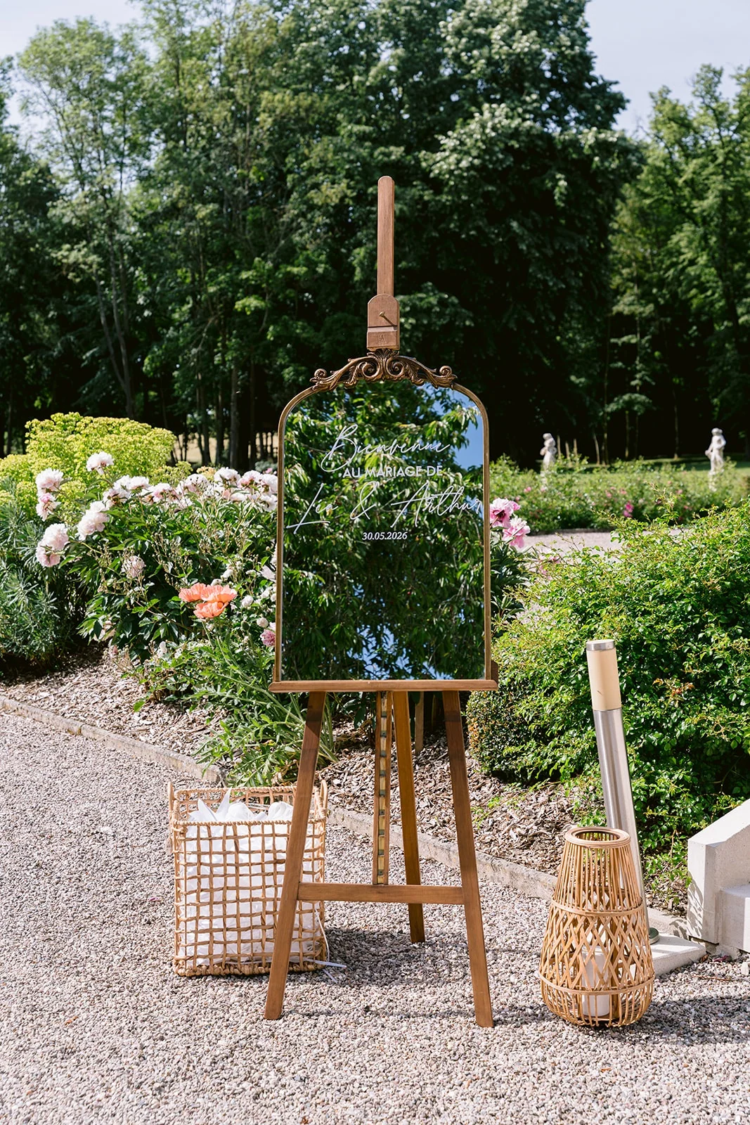 Un miroir décoré de fleurs repose sur un chevalet en bois, entre des lanternes et les massifs d’un jardin.