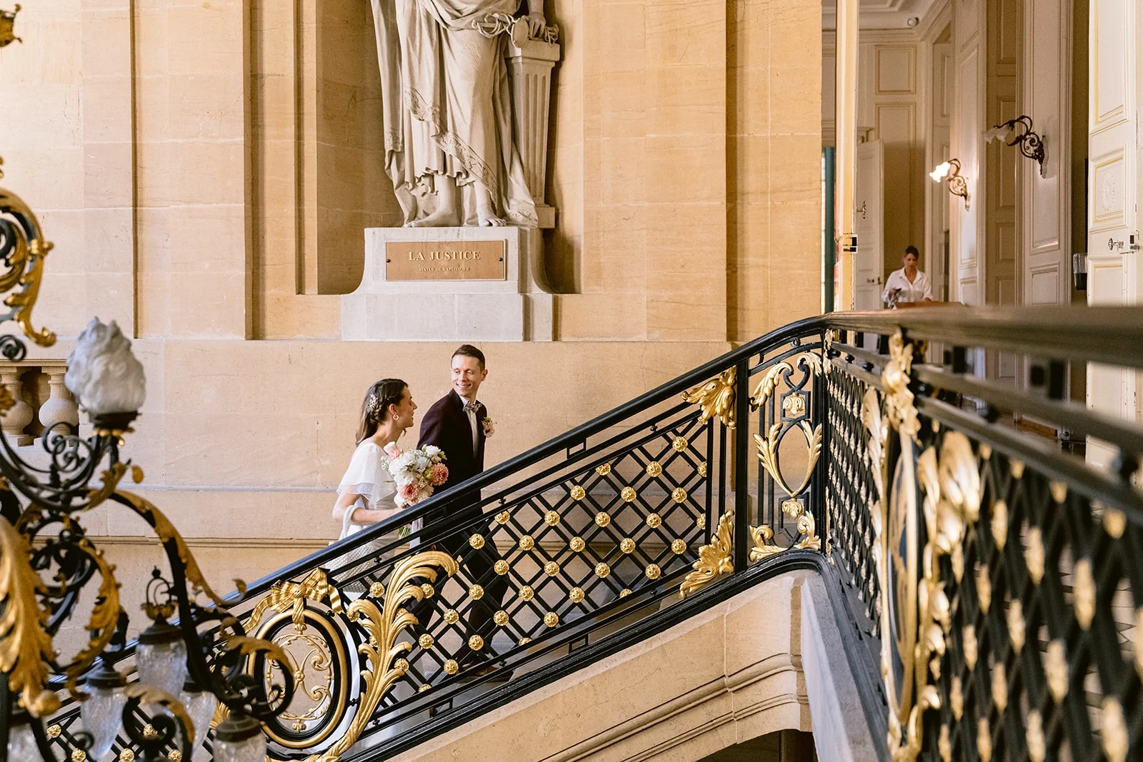Les mariés montent un grand escalier de pierre à la rampe noire et dorée, sous une statue.