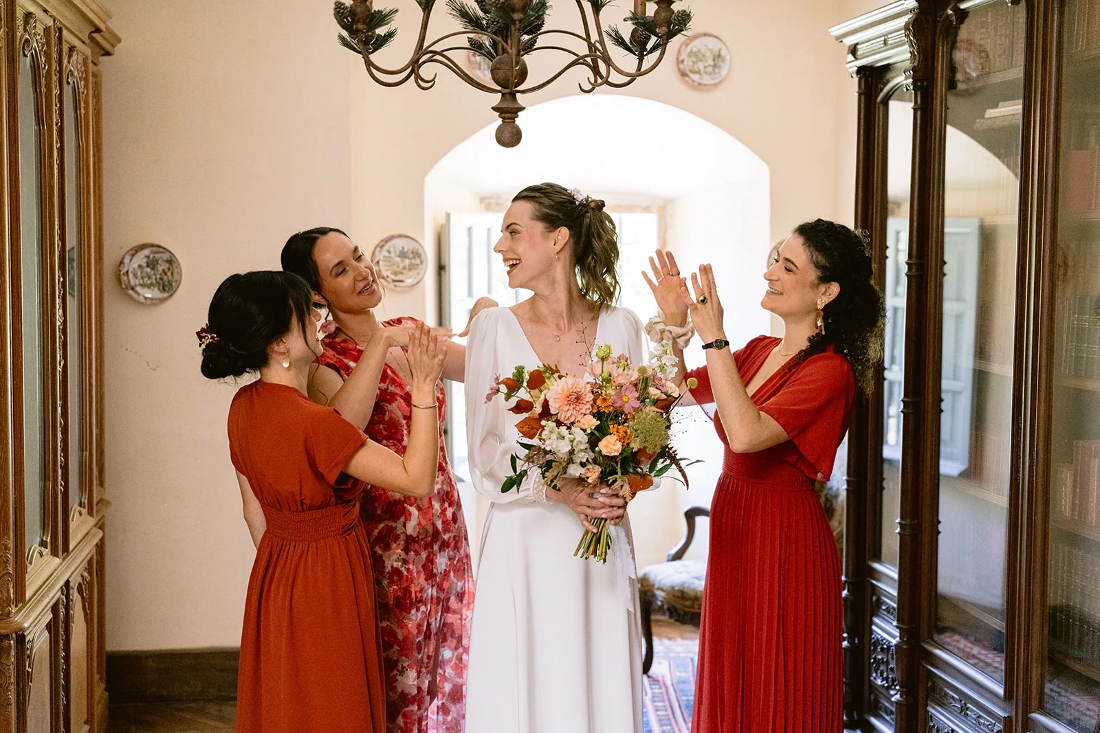 La mariée tient son bouquet coloré et rit avec trois femmes en robes rouges et orangées.
