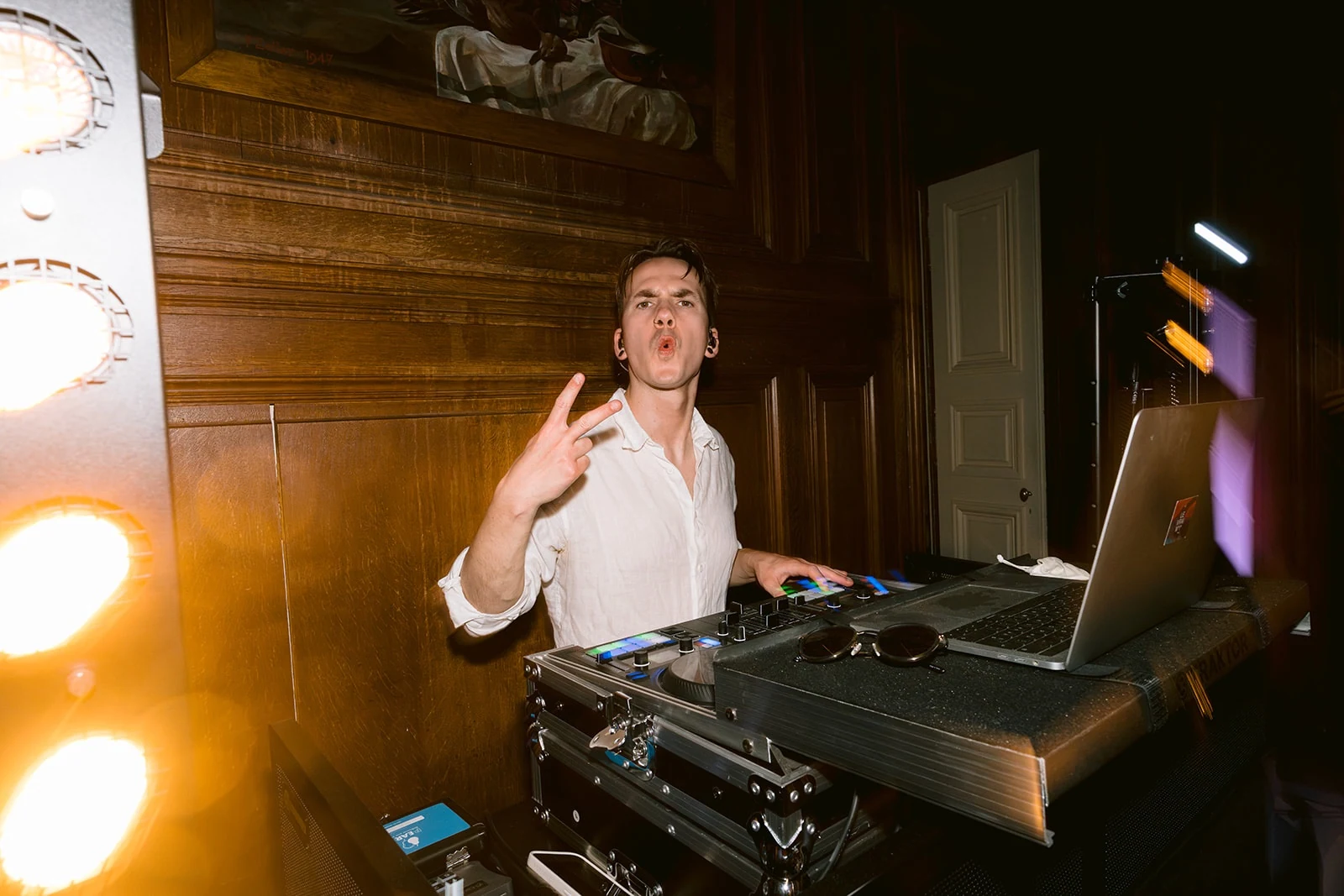 Un DJ en chemise blanche fait un geste de la main derrière ses platines et son ordinateur, près de projecteurs allumés.