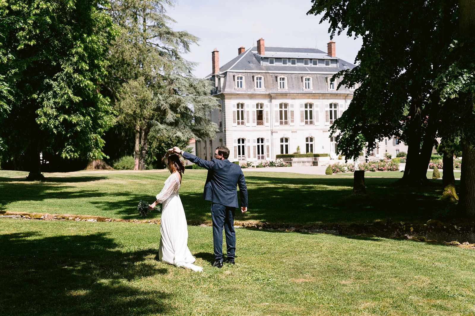 Le marié fait tourner la mariée par la main sur une pelouse, devant une grande demeure.