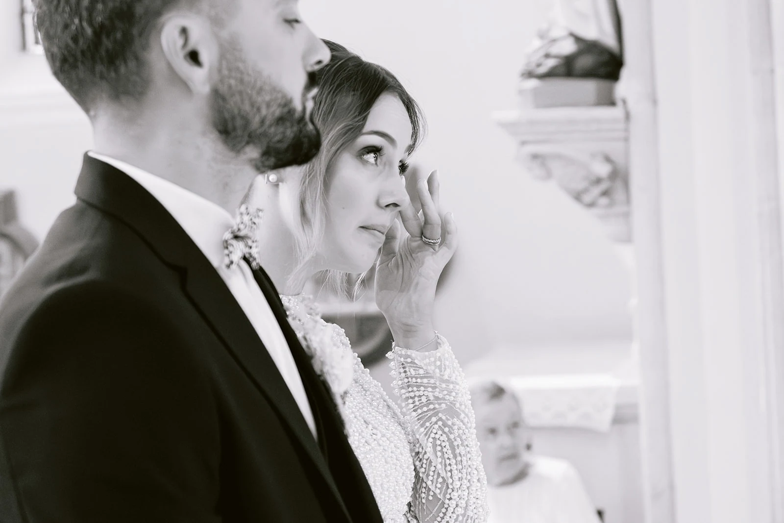 En noir et blanc, la mariée essuie une larme aux côtés du marié pendant la cérémonie.