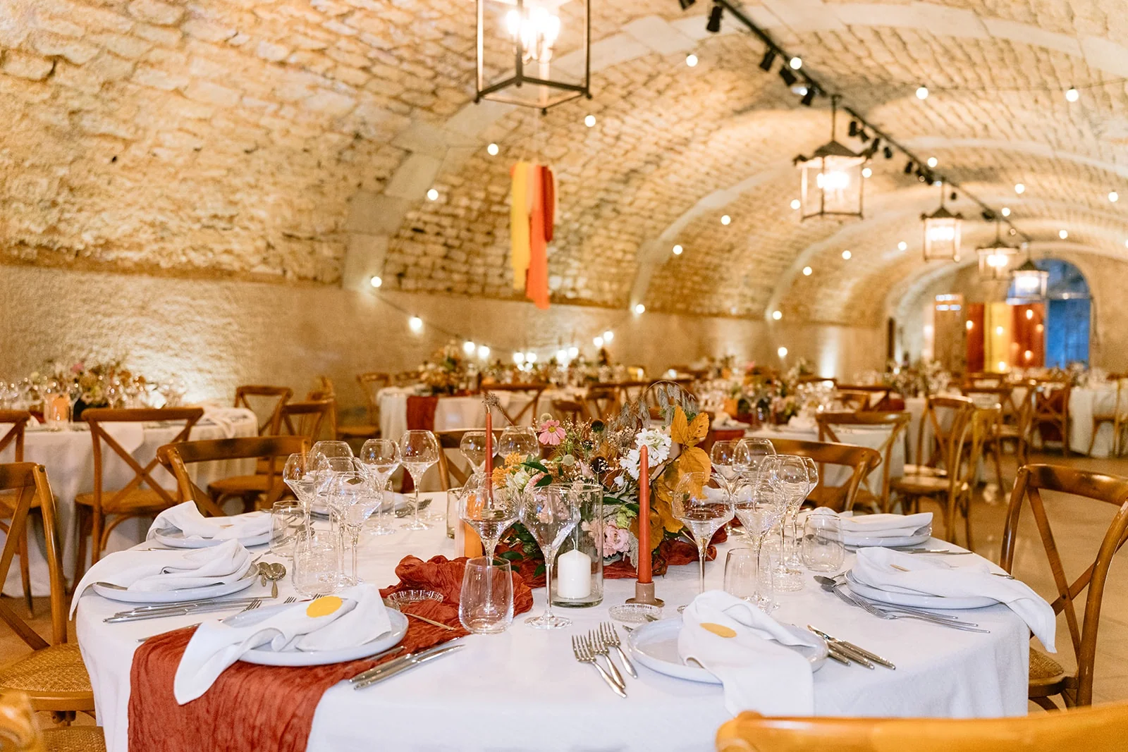 Une table ronde est dressée avec des fleurs, des bougies et un chemin de table orangé dans une salle voûtée.