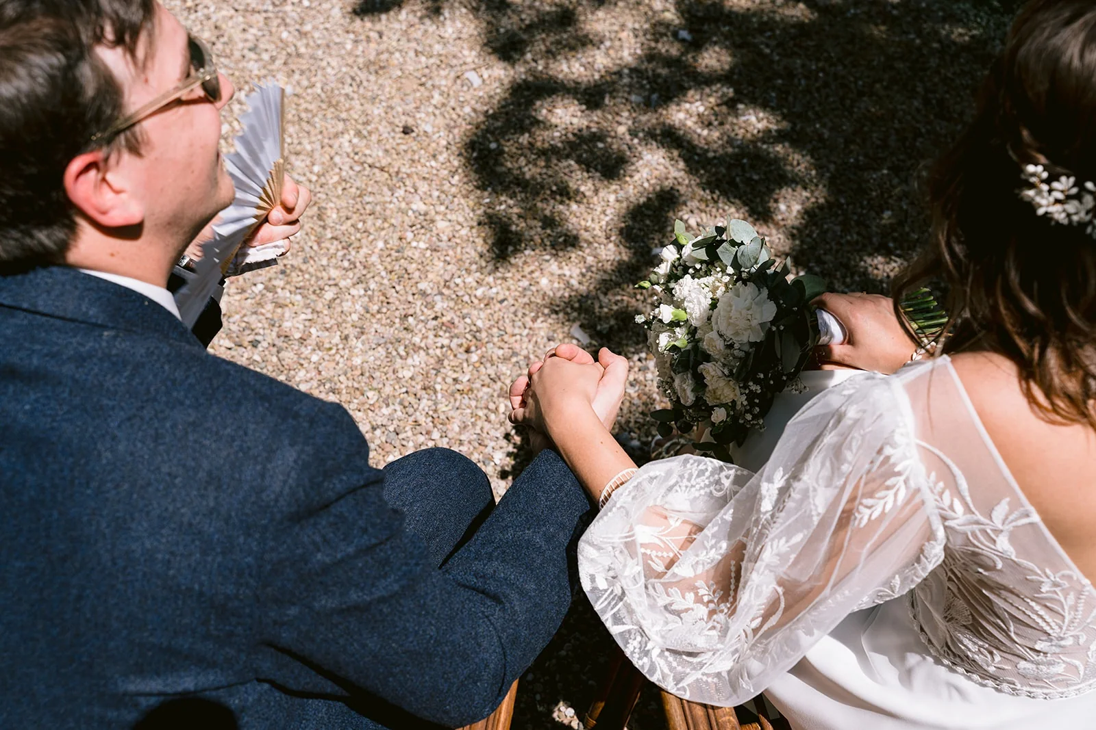 Vue de dos des mariés assis côte à côte pendant la cérémonie en plein air, le bouquet posé entre eux.