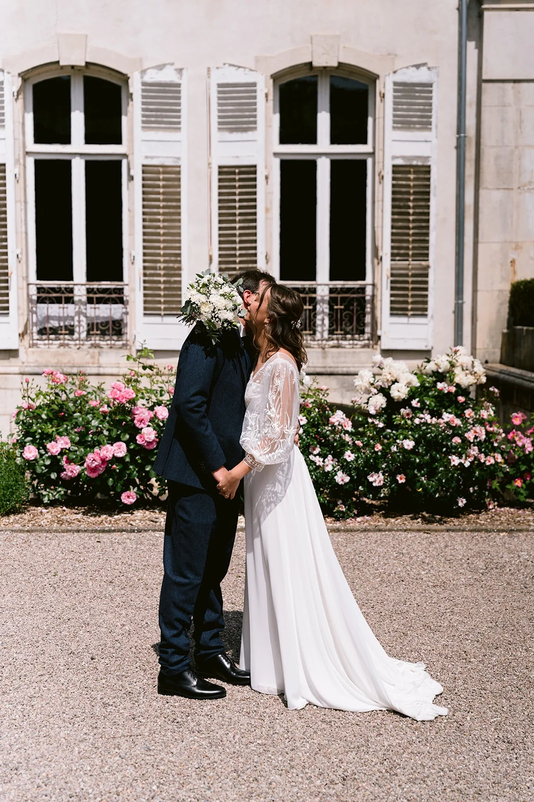 Les mariés s’embrassent devant une façade et des rosiers, leur visage partiellement caché par le bouquet blanc.