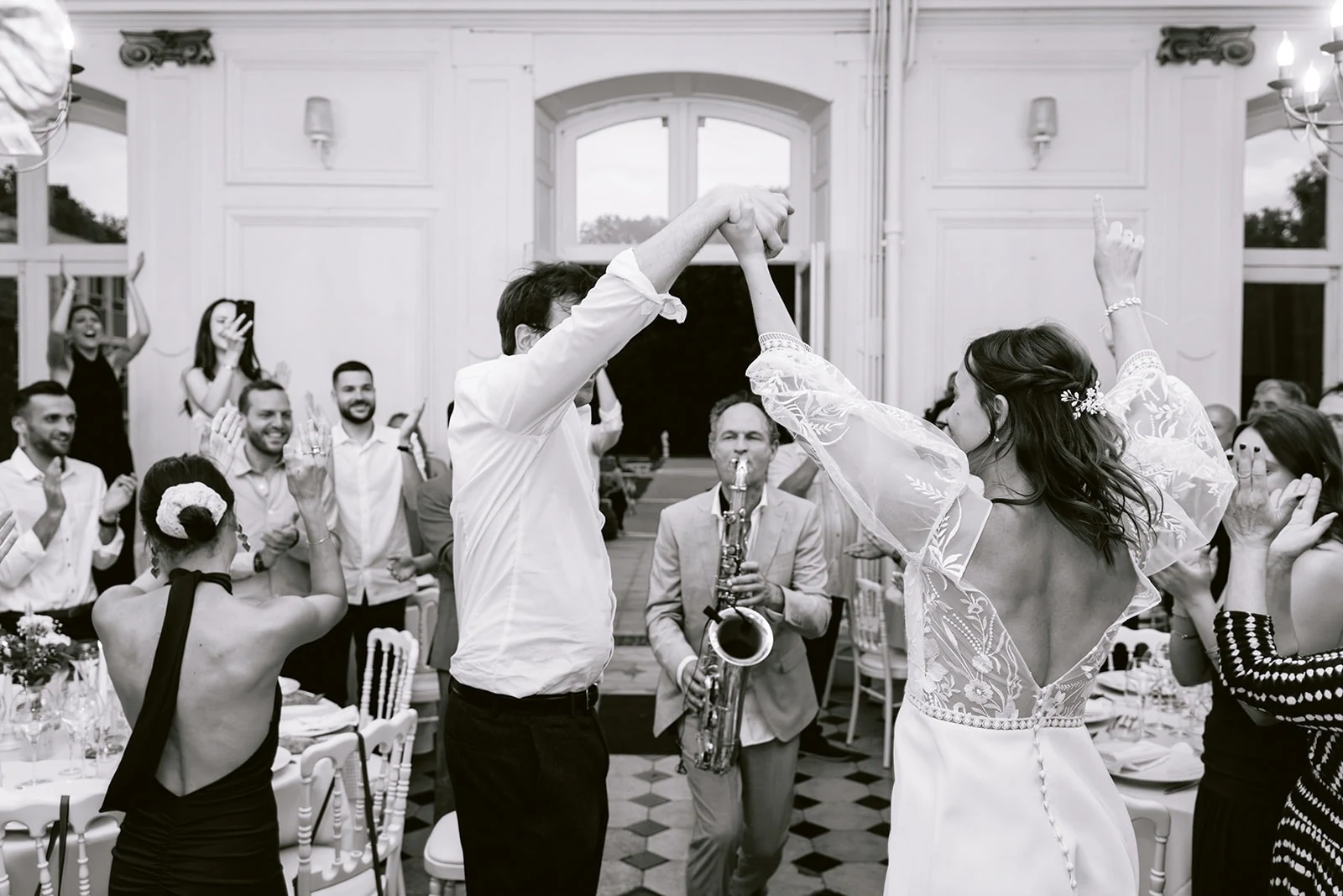 En noir et blanc, les mariés dansent les bras levés devant un saxophoniste et leurs invités.