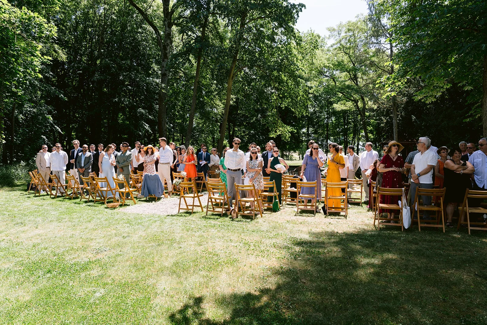 Les invités se tiennent debout devant des rangées de chaises en bois, dans une clairière ombragée.
