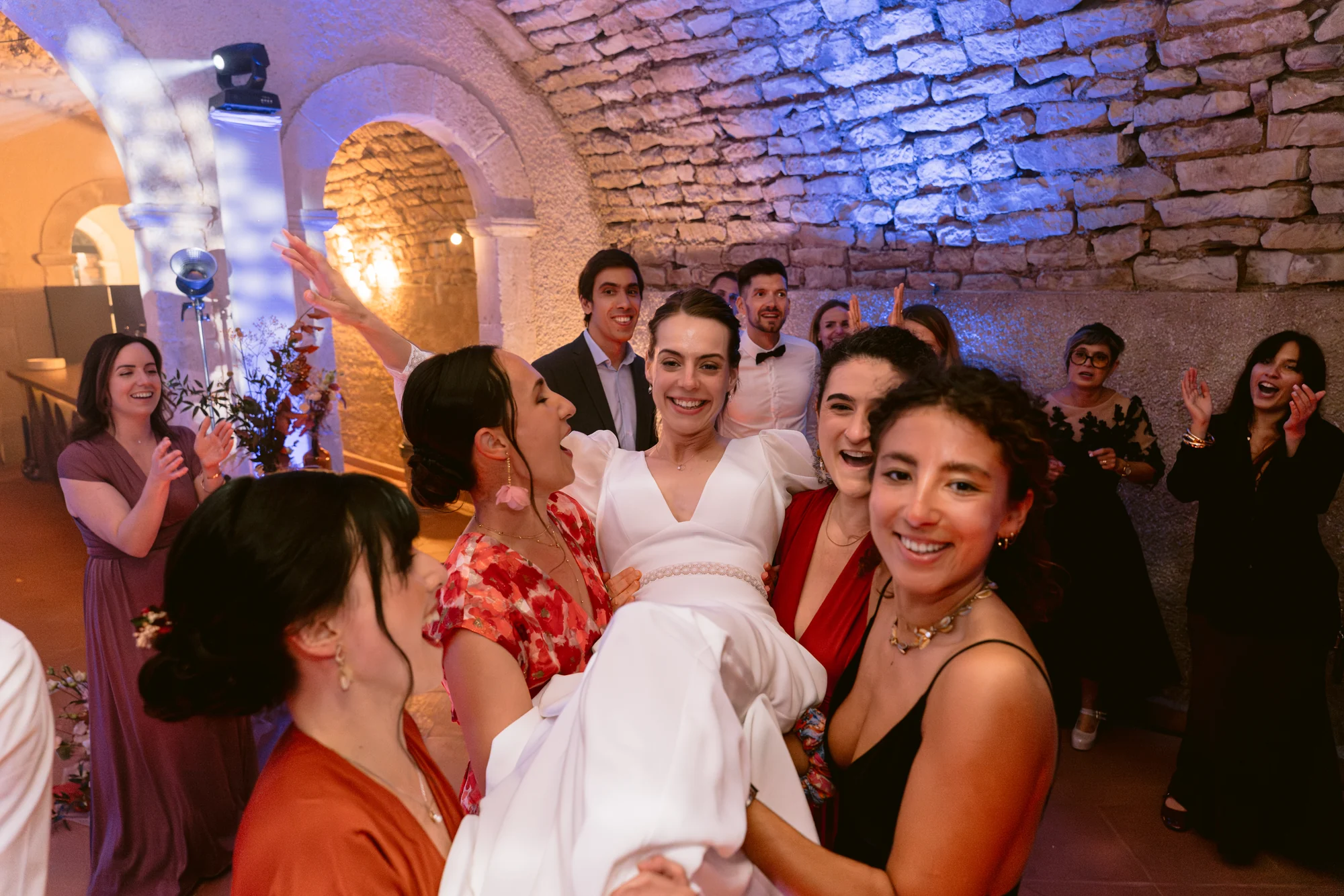 Des amies portent la mariée au milieu de la fête, sous des voûtes en pierre.