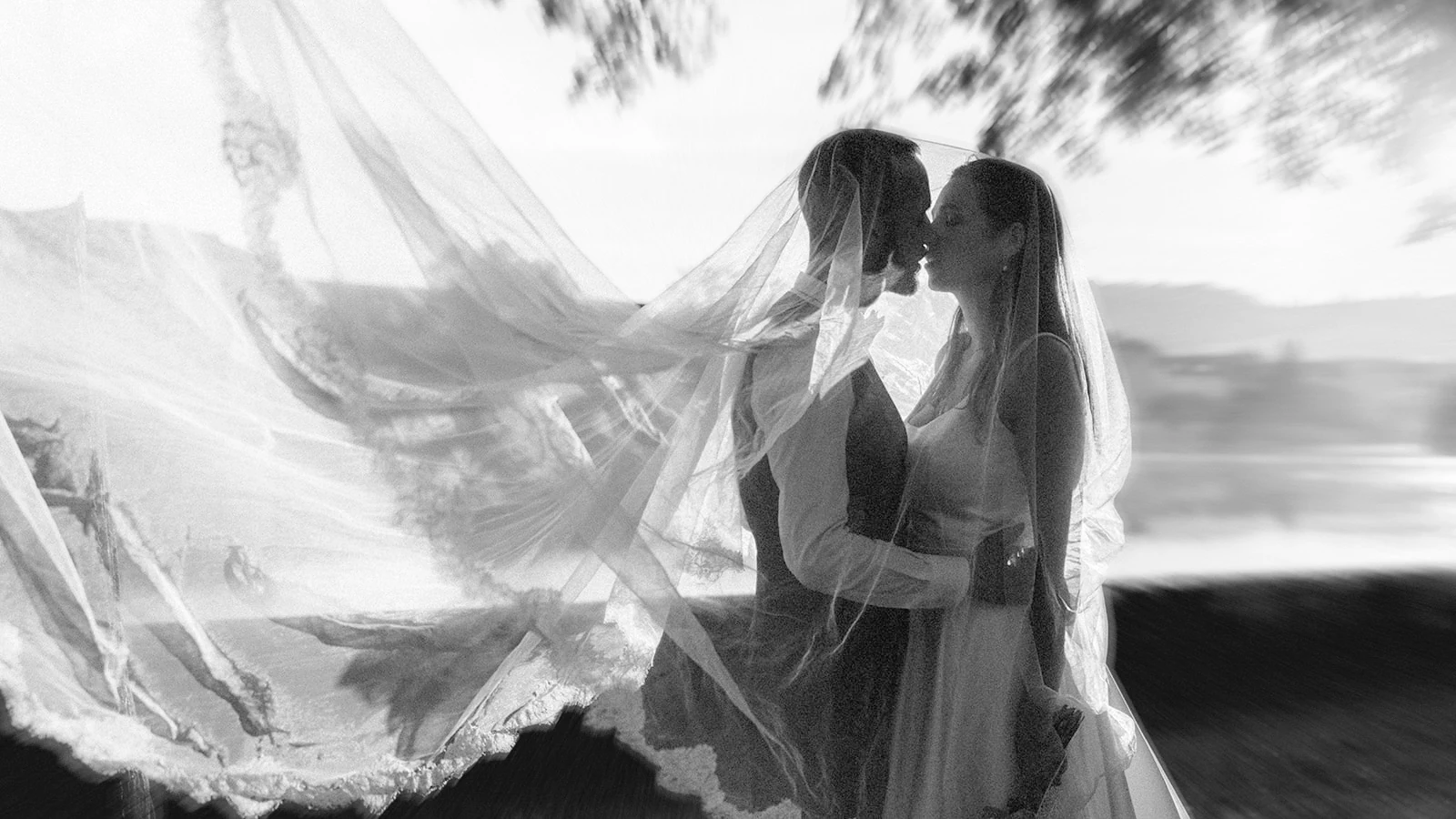 En noir et blanc, les mariés s’embrassent sous un voile soulevé par le vent.