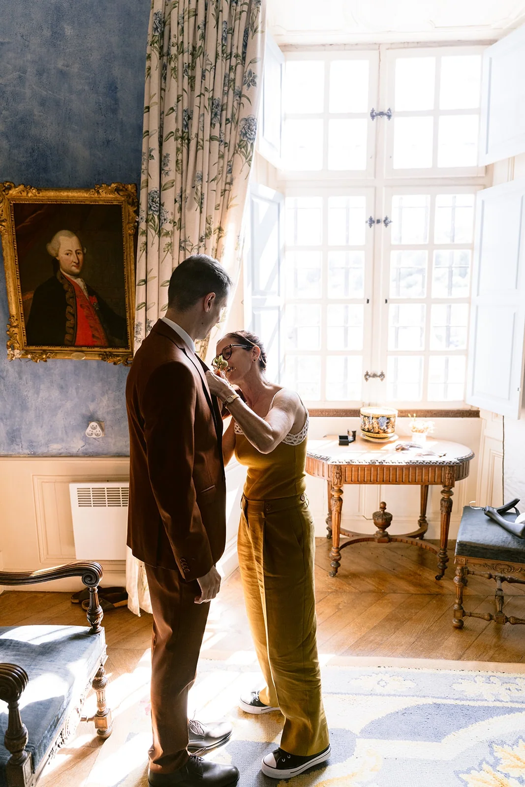 Une femme en robe ocre ajuste la boutonnière du marié devant une grande fenêtre.
