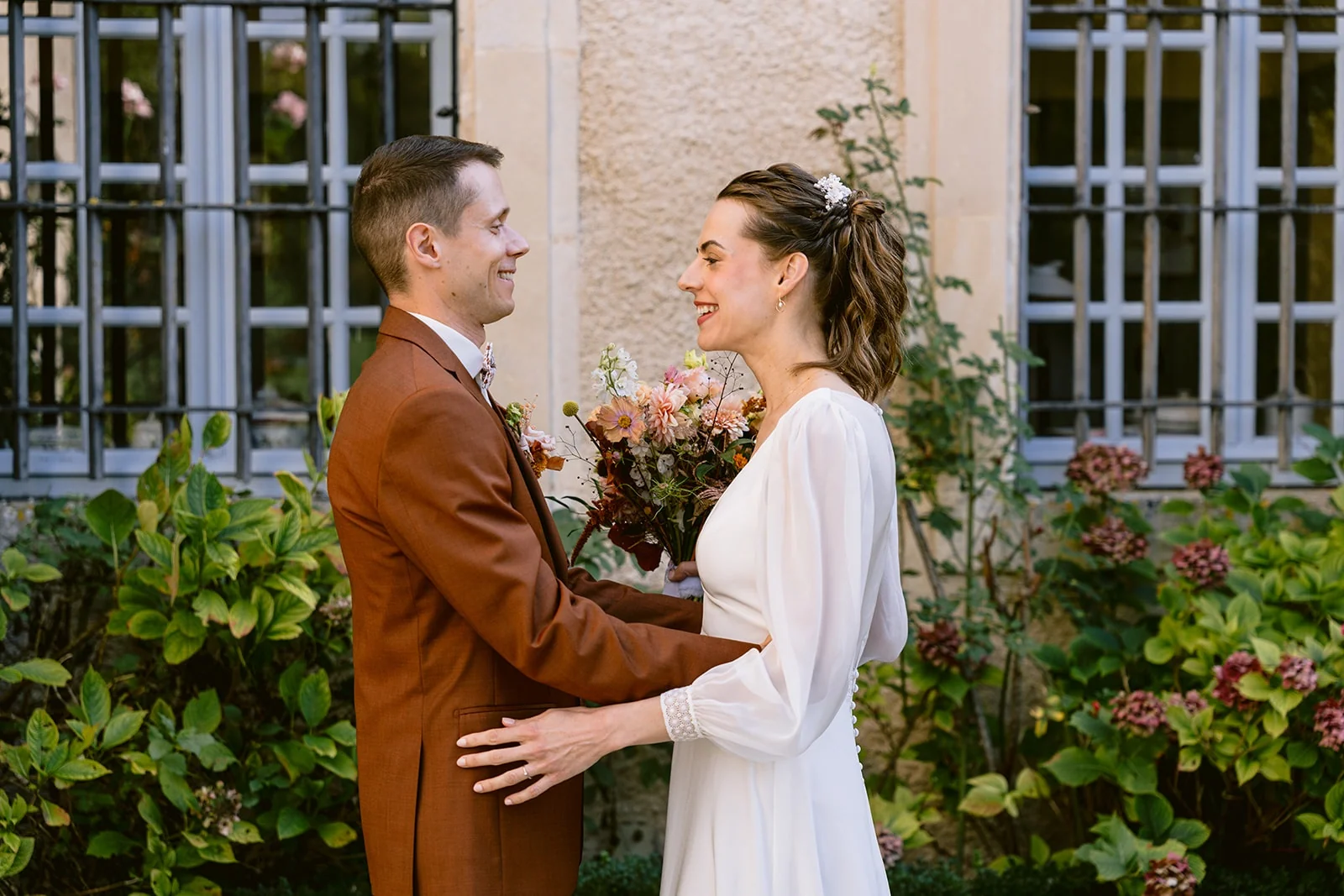 Les mariés se regardent en souriant, les mains posées sur les bras l’un de l’autre, dans une cour fleurie.