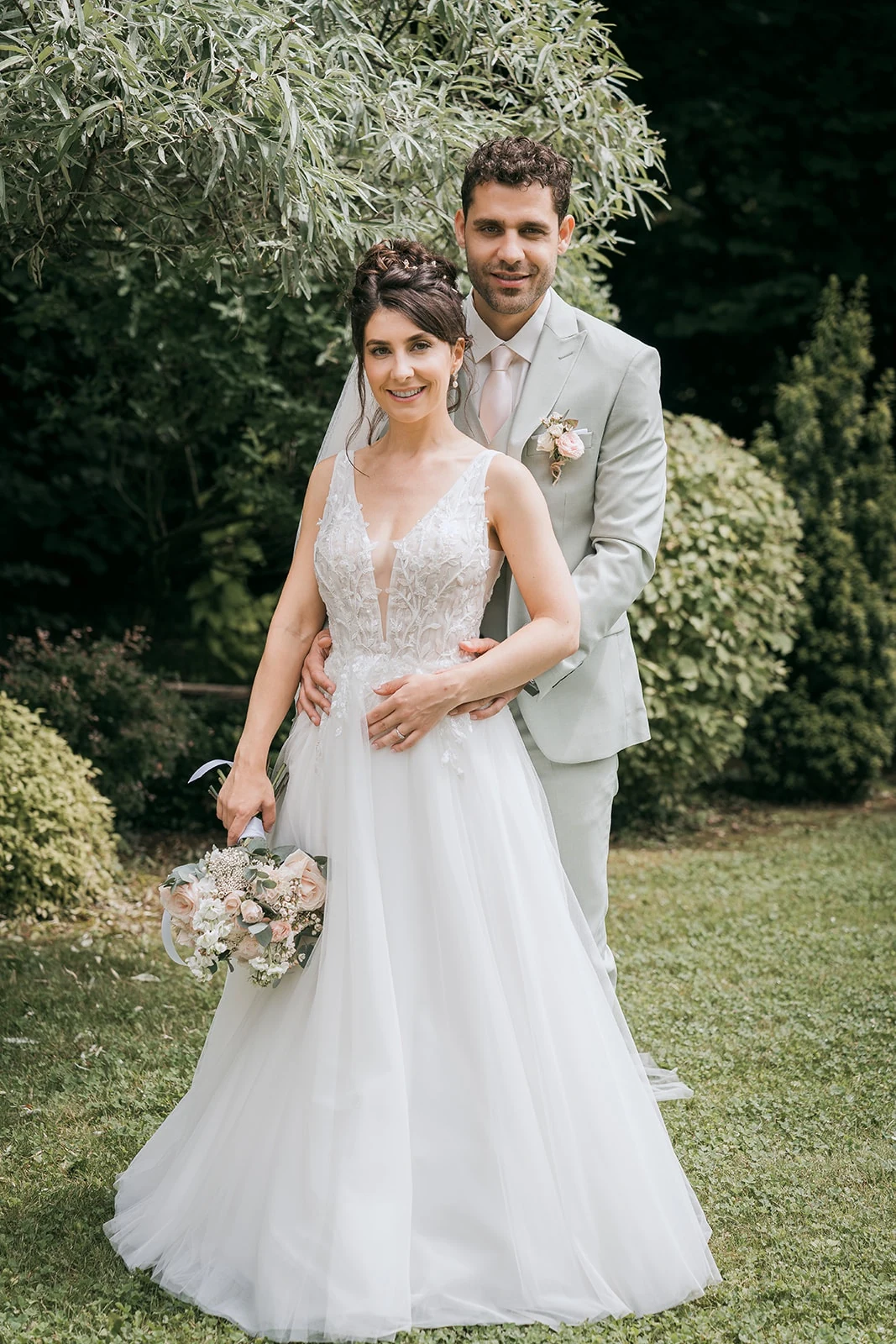 Portrait des mariés dans un jardin, la mariée tenant son bouquet devant le marié en costume clair.