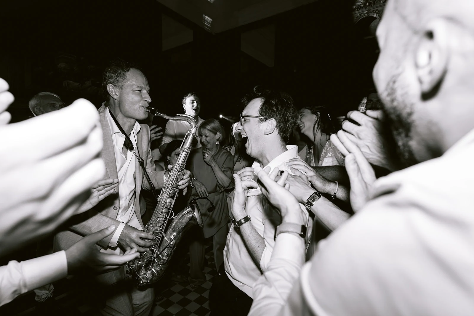 En noir et blanc, un saxophoniste joue face au marié souriant, entouré d’invités qui applaudissent.